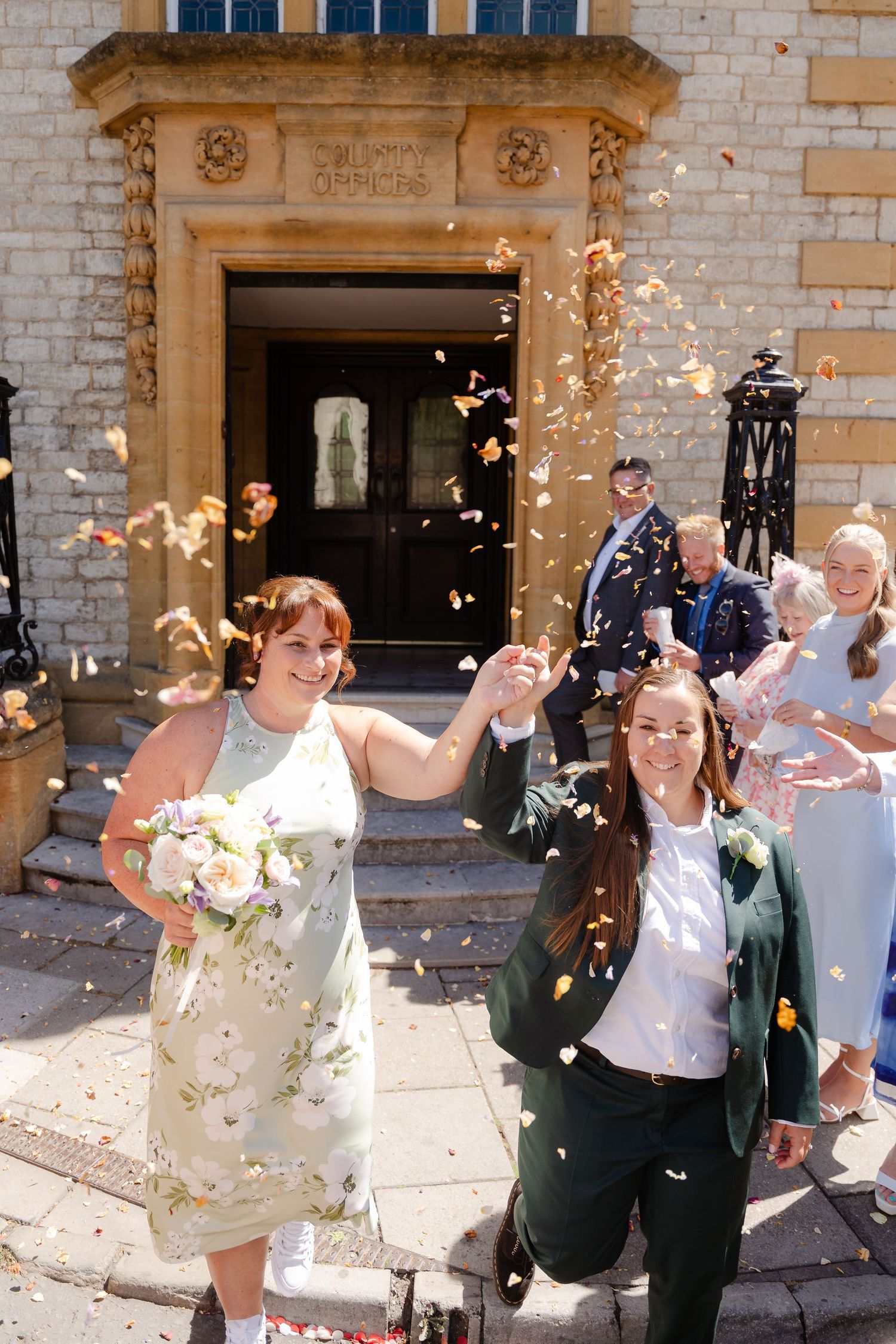 Two brides celebrating outside a wedding venue with confetti being thrown by guests on sunny day.