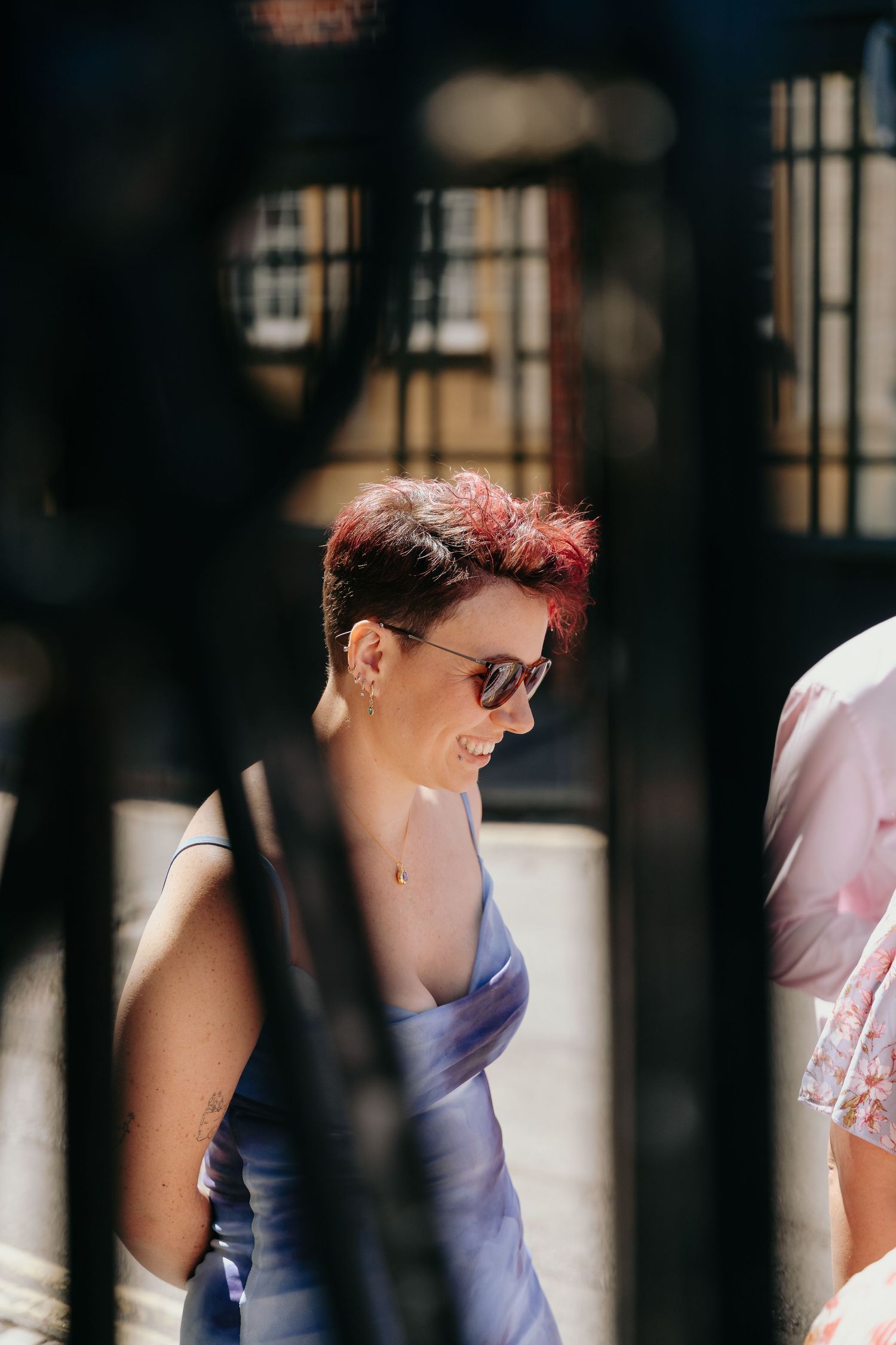 Smiling woman with short pink hair and sunglasses wearing a blue dress, photographed through metal bars in sunlight.