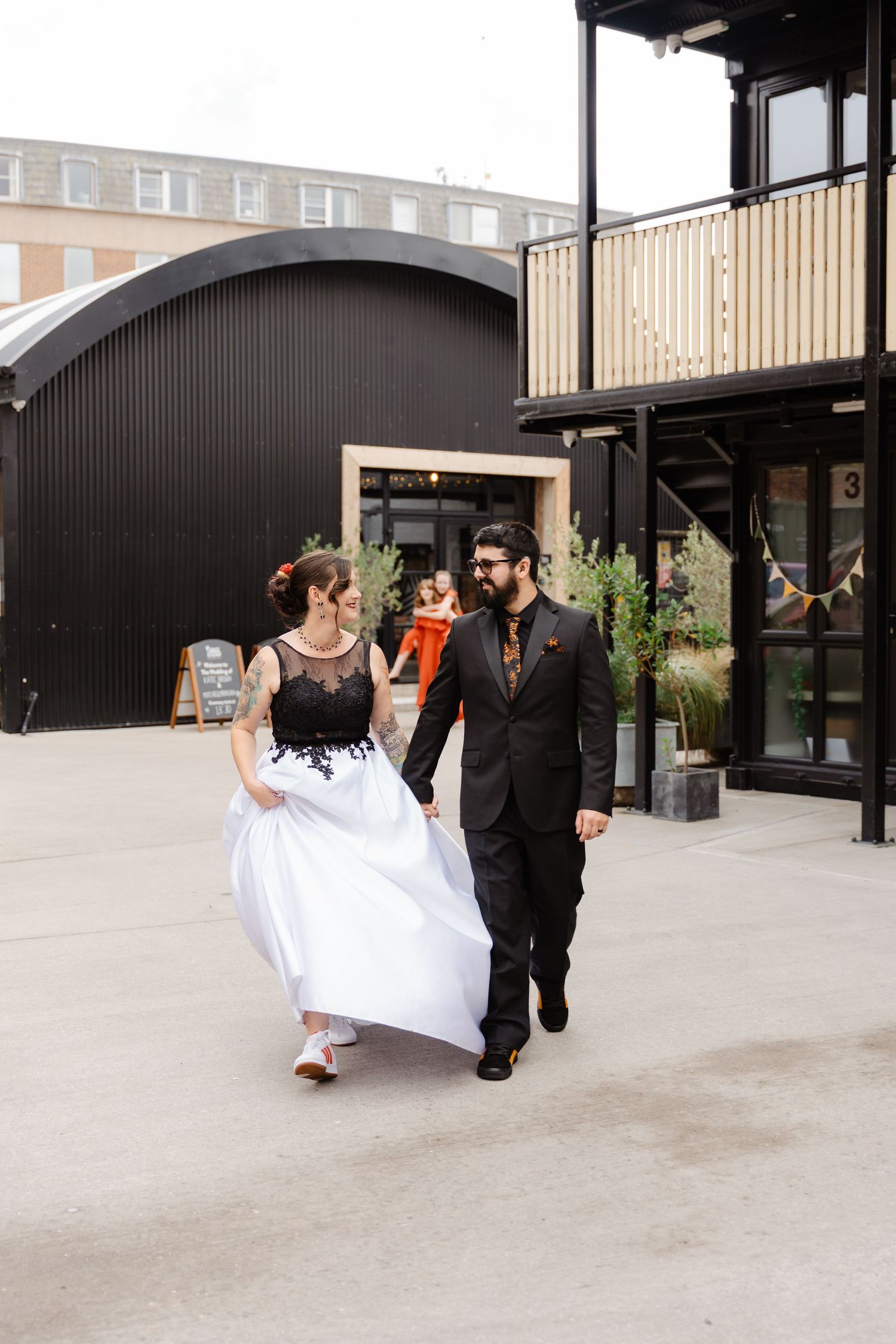 Bride in black and white wedding dress walks hand in hand with groom in black suit at outdoor industrial venue.