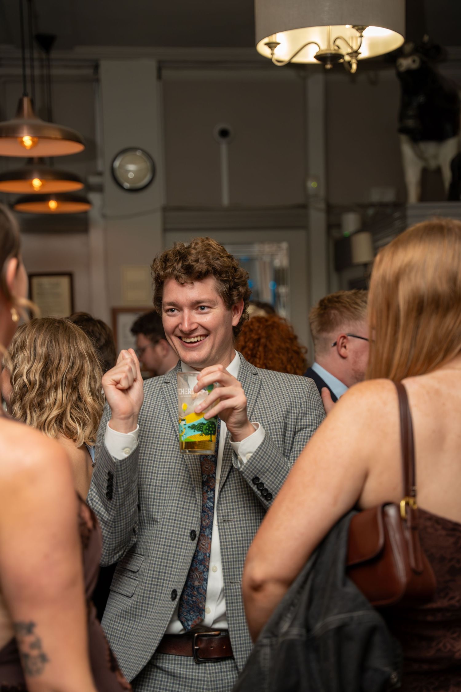 A smiling man in a gray plaid suit holds a drink at a lively indoor social gathering or party event.