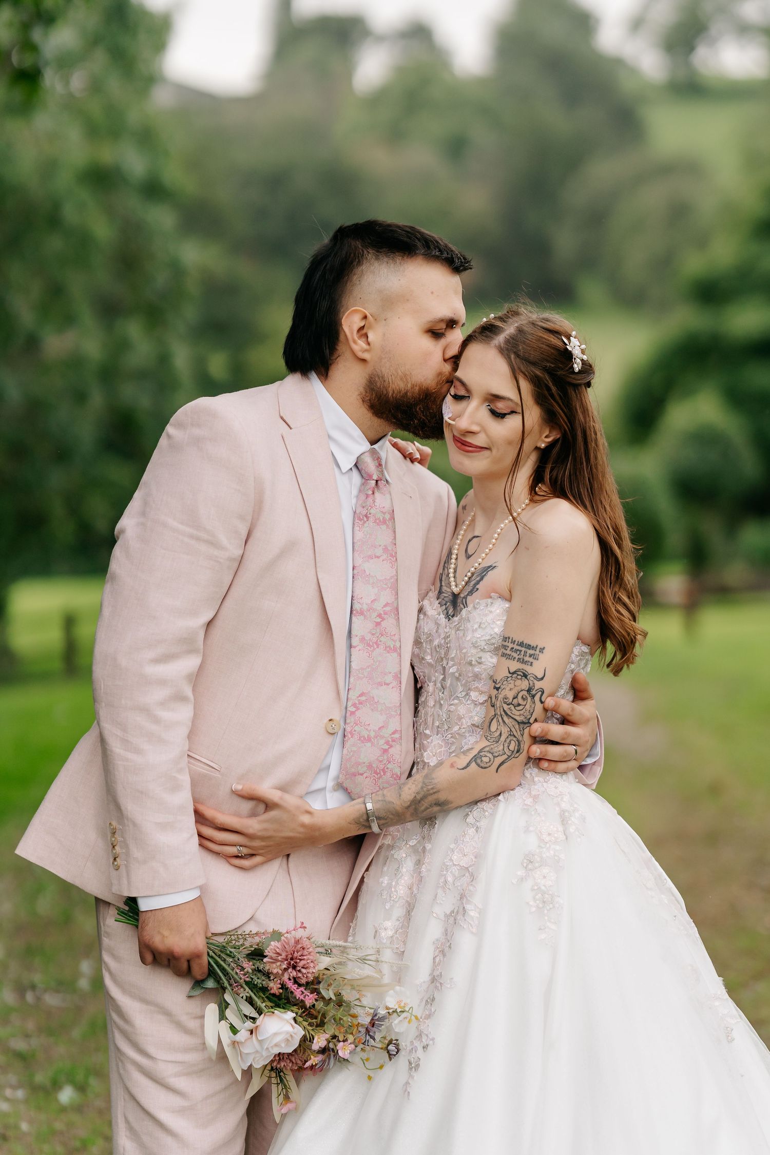 Groom in pink suit kisses bride's forehead outdoors, she holds a wildflower bouquet in a lace wedding gown.