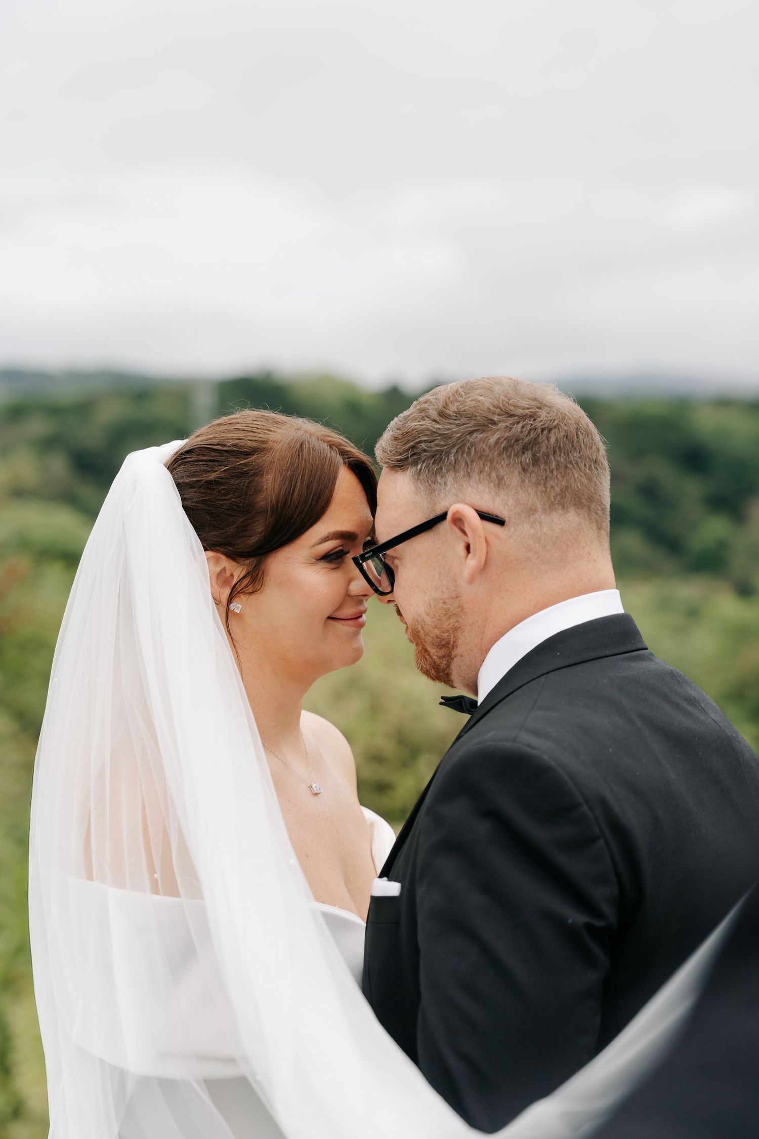 Bride and groom share an intimate moment outdoors, with her white veil flowing and lush greenery in the background.