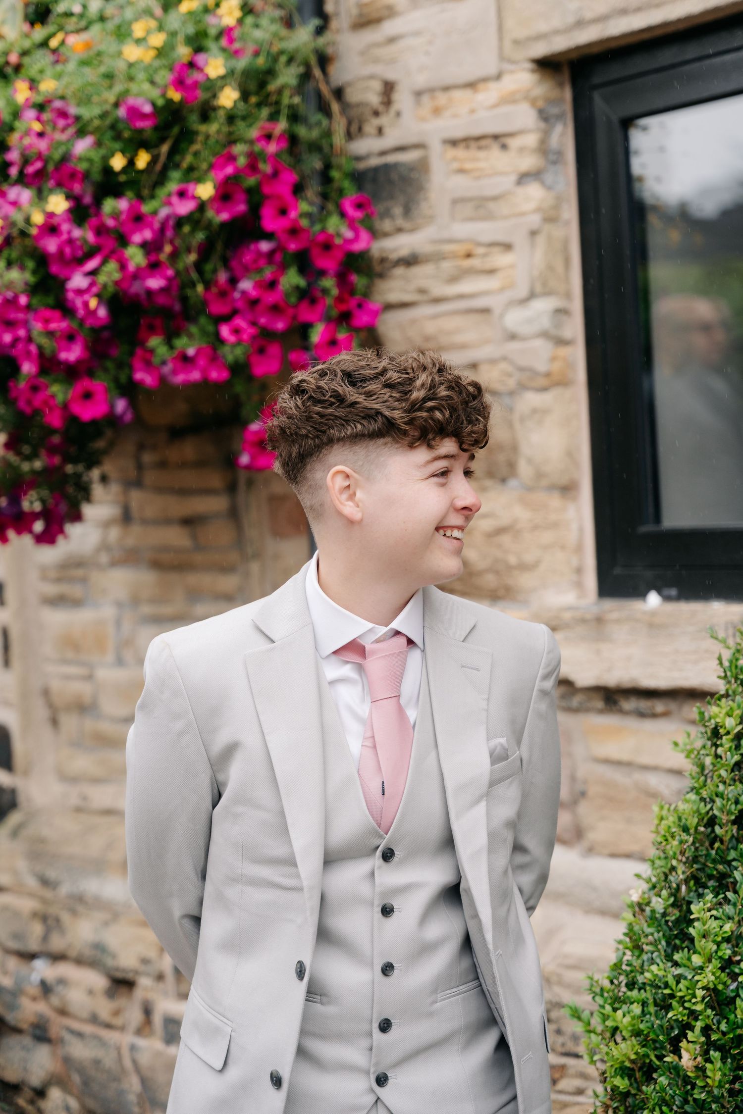 Young man in light gray three-piece suit with pink tie smiling outdoors near stone wall with pink flowers.
