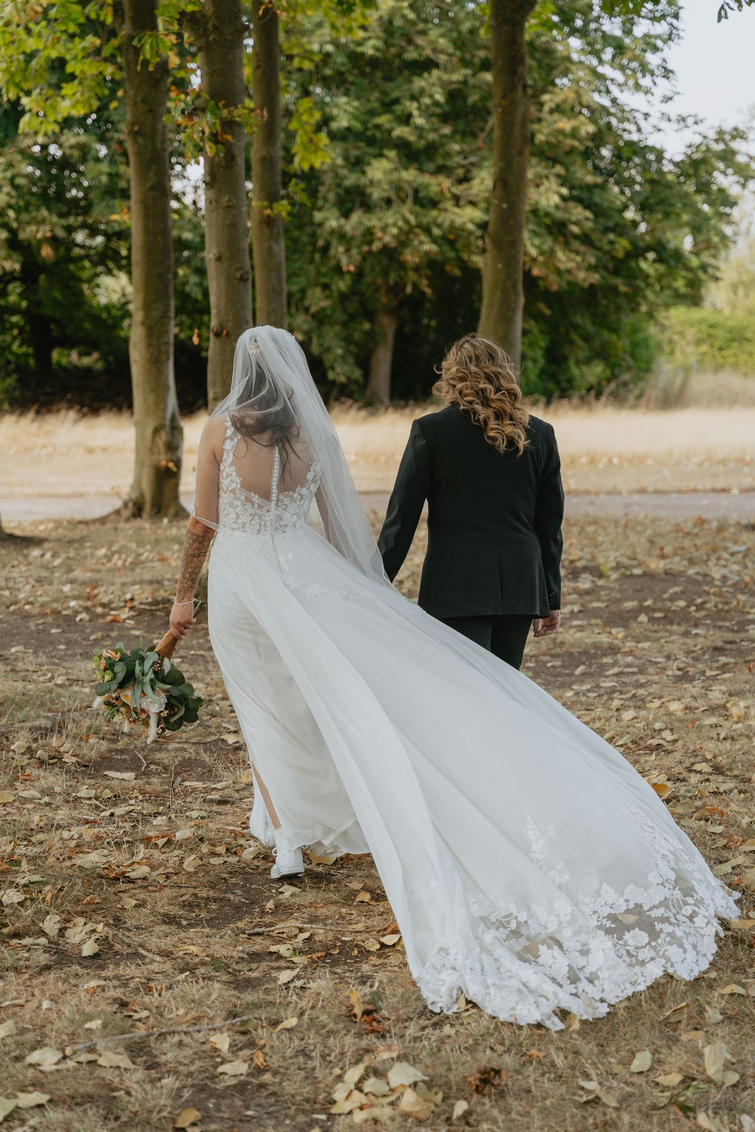 Two brides walk hand-in-hand through a tree-lined path, one in a flowing lace wedding gown with veil and bouquet.
