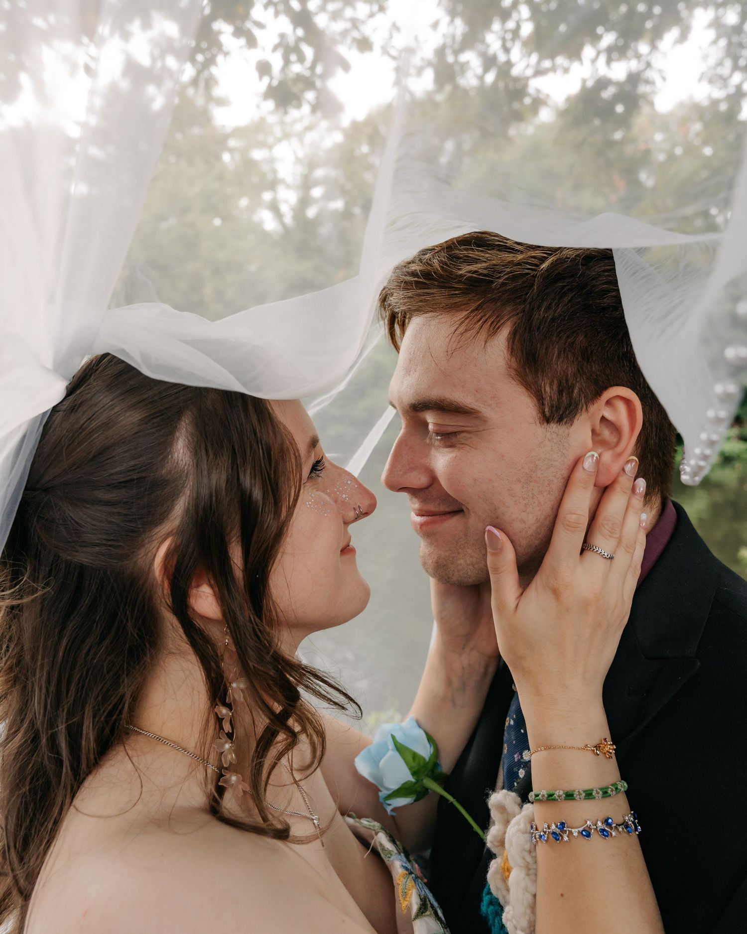 Bride and groom share a romantic moment under a wedding veil, with the bride holding a blue rose outdoors.