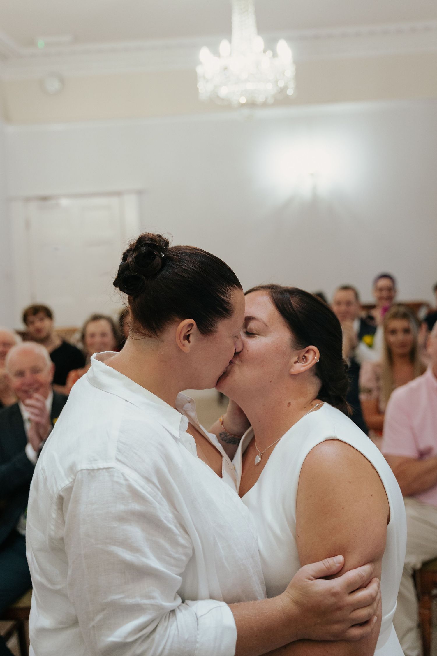 Two brides share a first kiss at their wedding ceremony, both wearing white, with guests celebrating in the background.