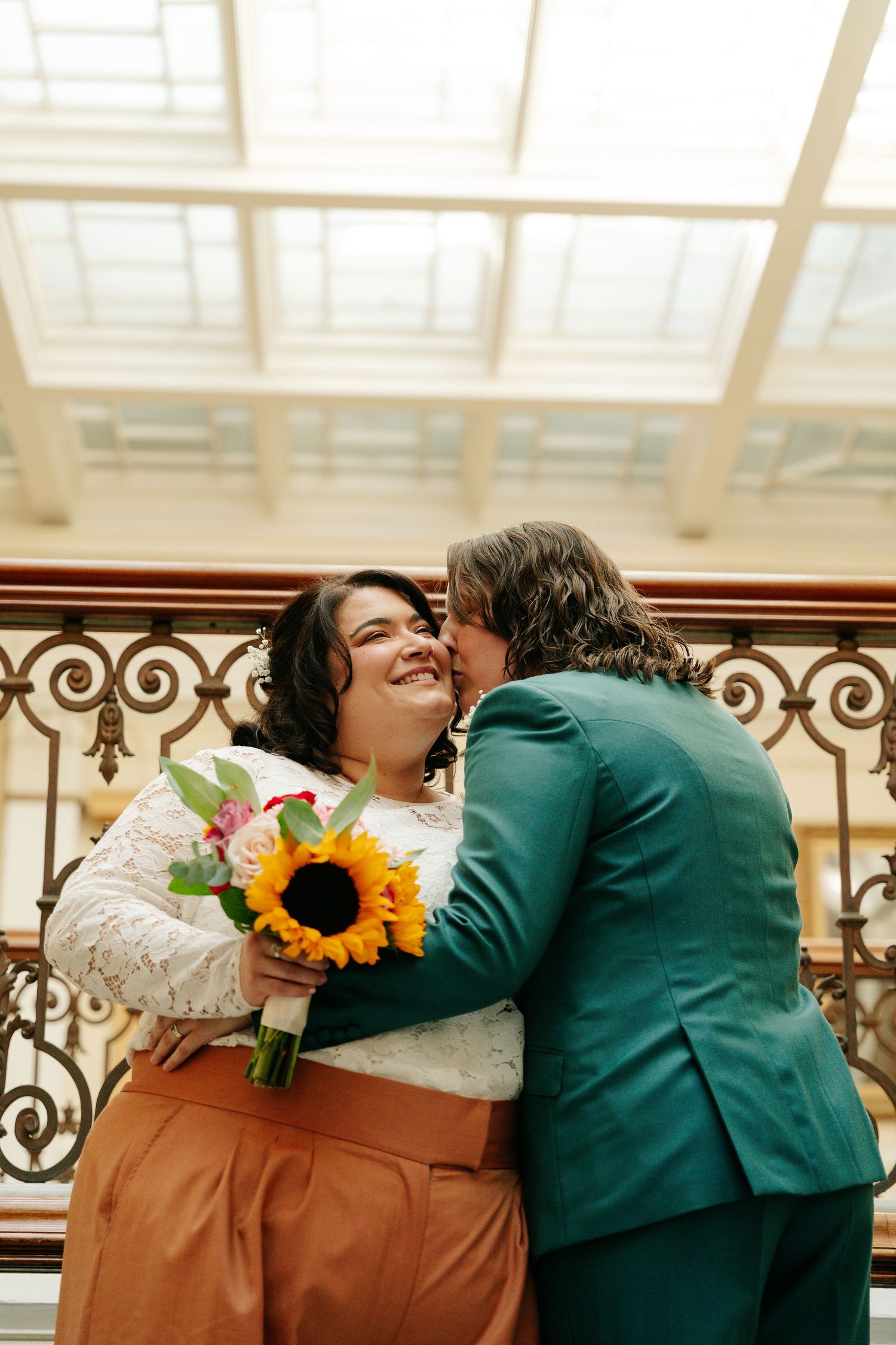 Bride holding sunflower bouquet smiles as groom in teal suit kisses her cheek on ornate balcony.