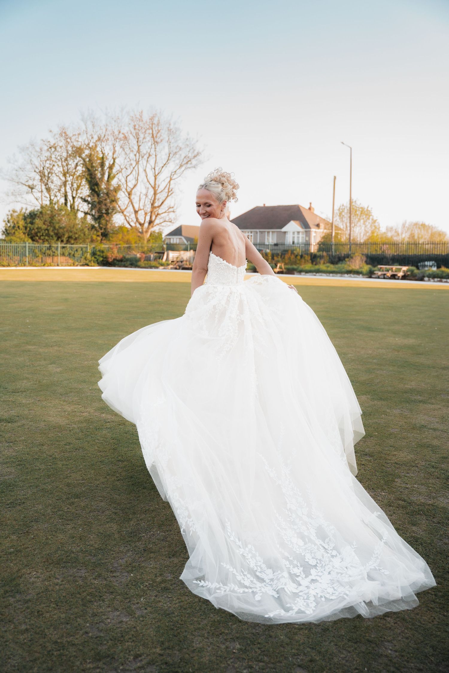 Bride in a flowing white lace ball gown twirling on a green lawn at golden hour.