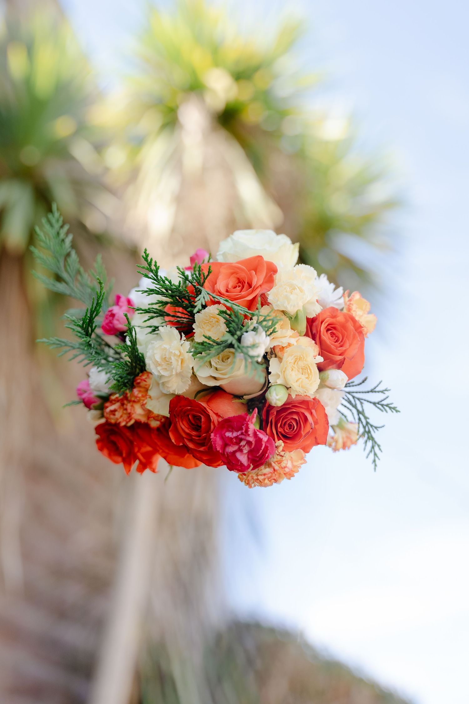 Vibrant wedding bouquet with orange, red, and cream roses with green foliage against a blue sky backdrop.
