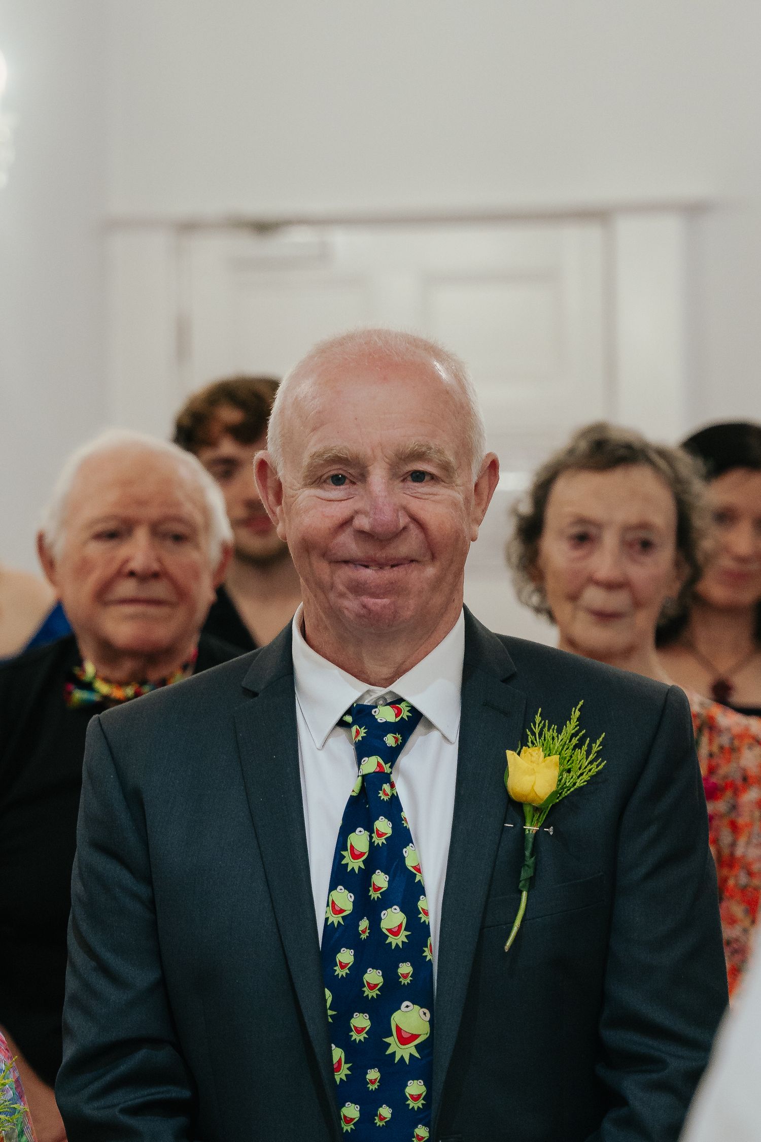 Smiling bald man in navy suit with Kermit the Frog tie and yellow rose boutonniere at a wedding ceremony.