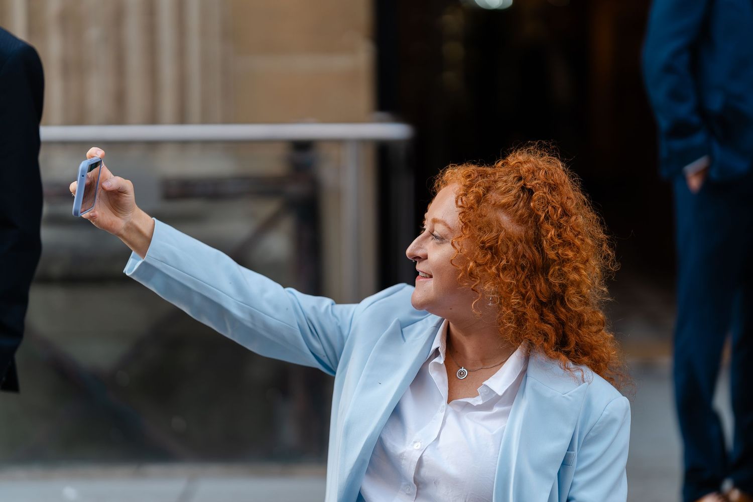 Red-haired woman in light blue blazer taking a selfie outdoors, smiling confidently in an urban setting.