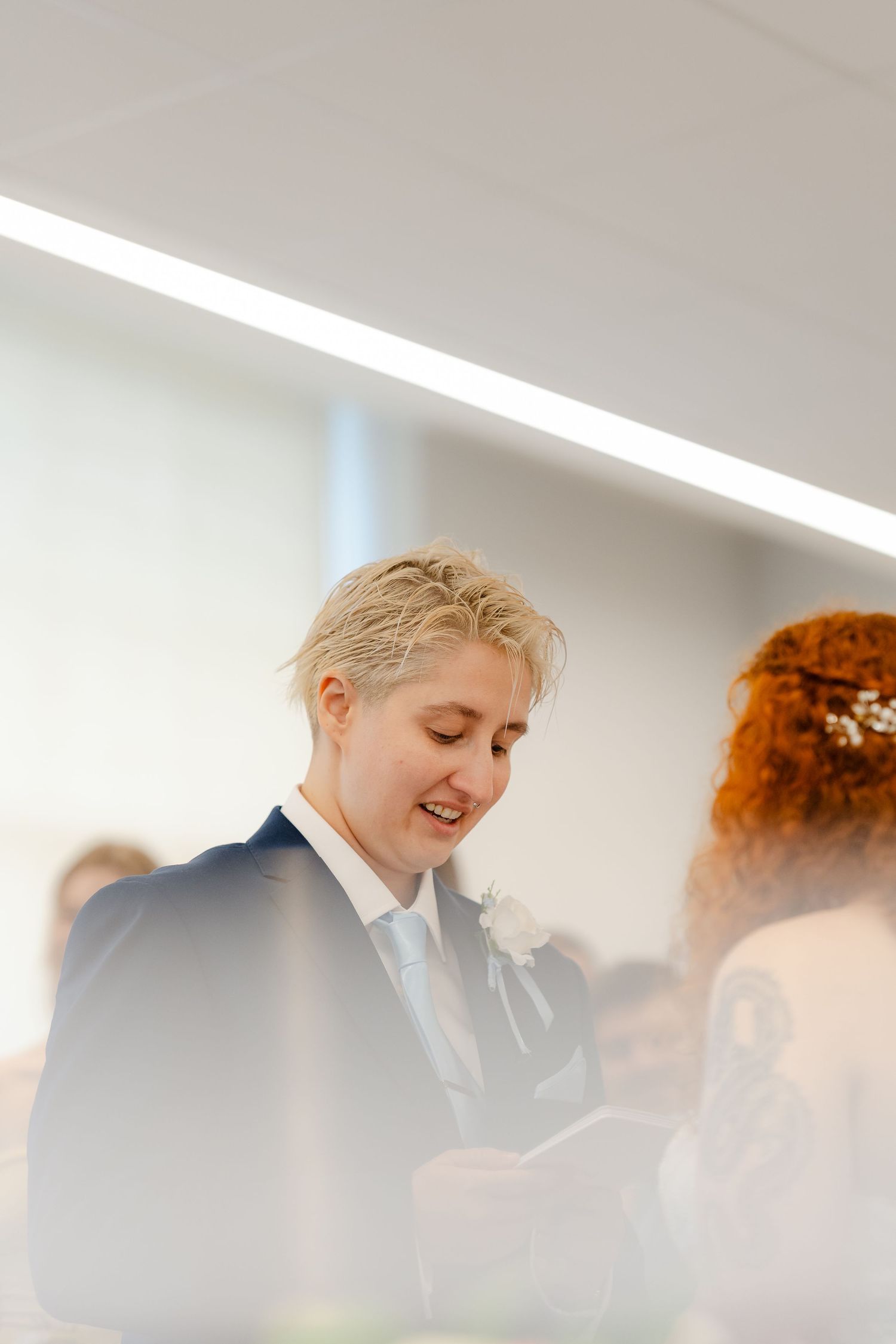 Smiling blonde person in navy suit with white boutonniere at a wedding ceremony, facing a redhead partner.
