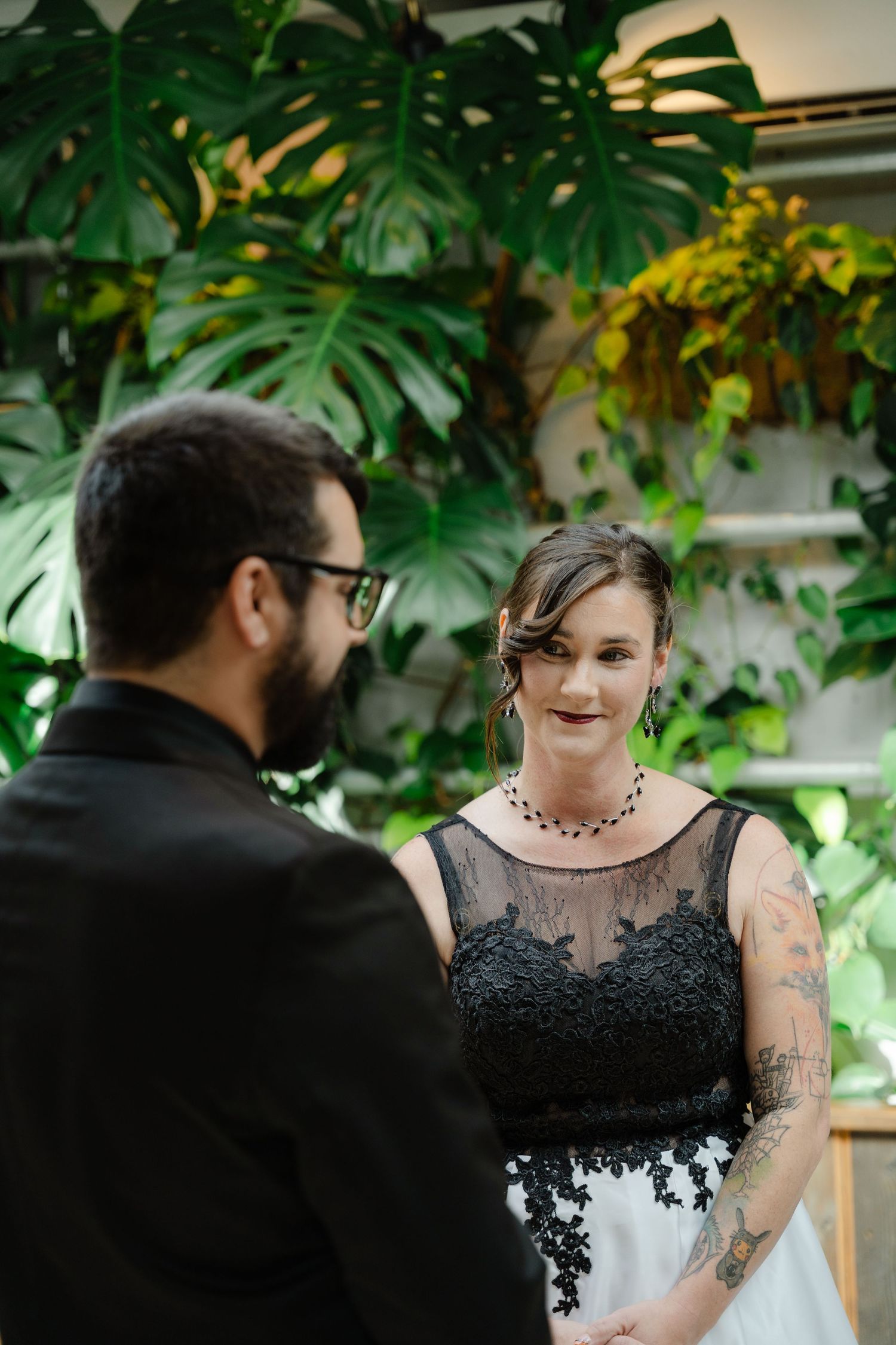 Bride in black lace and white wedding dress smiles at groom during ceremony with tropical monstera leaves backdrop.