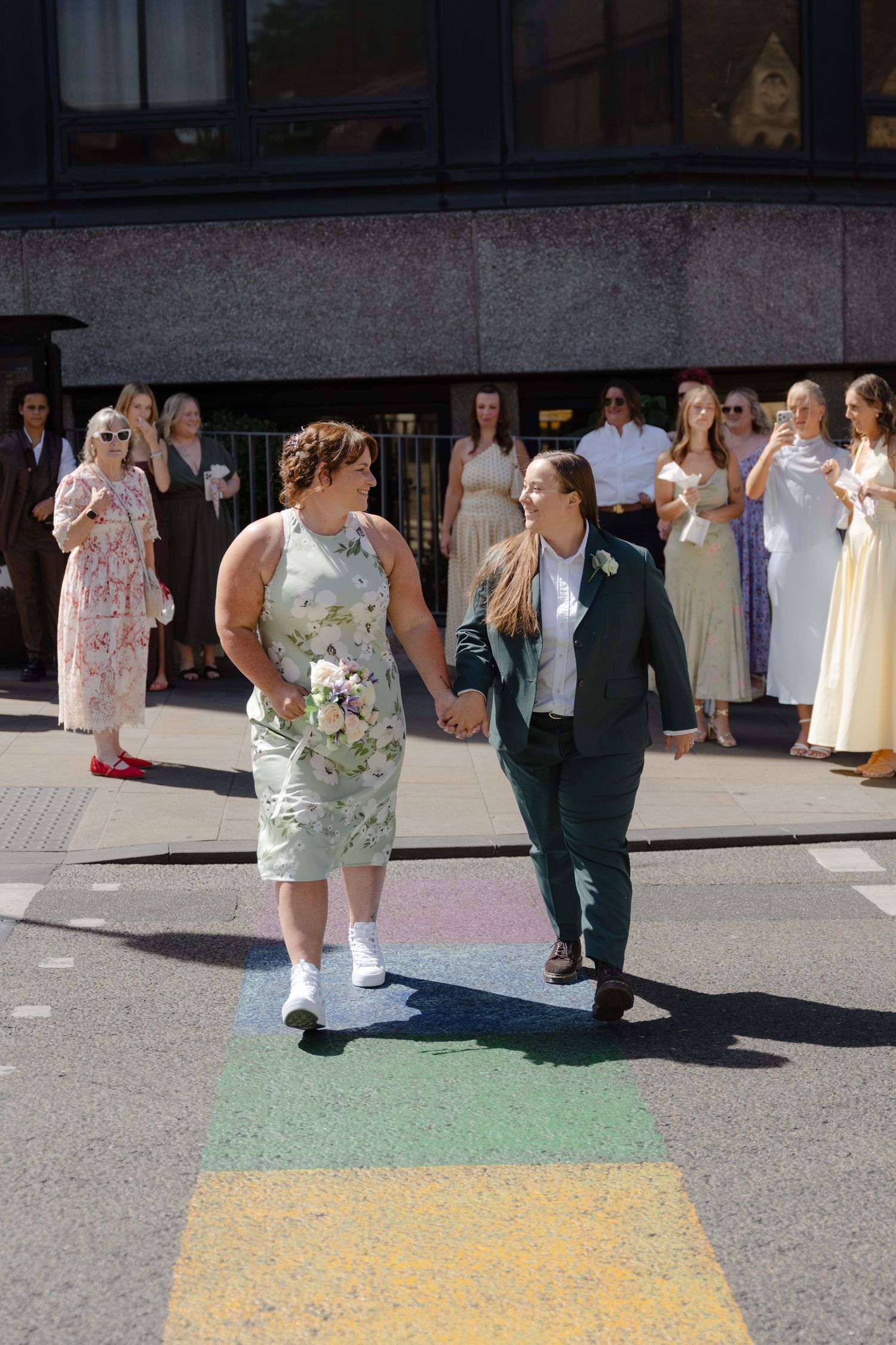 Two brides holding hands walk across a rainbow crosswalk, celebrated by guests on a sunny wedding day.