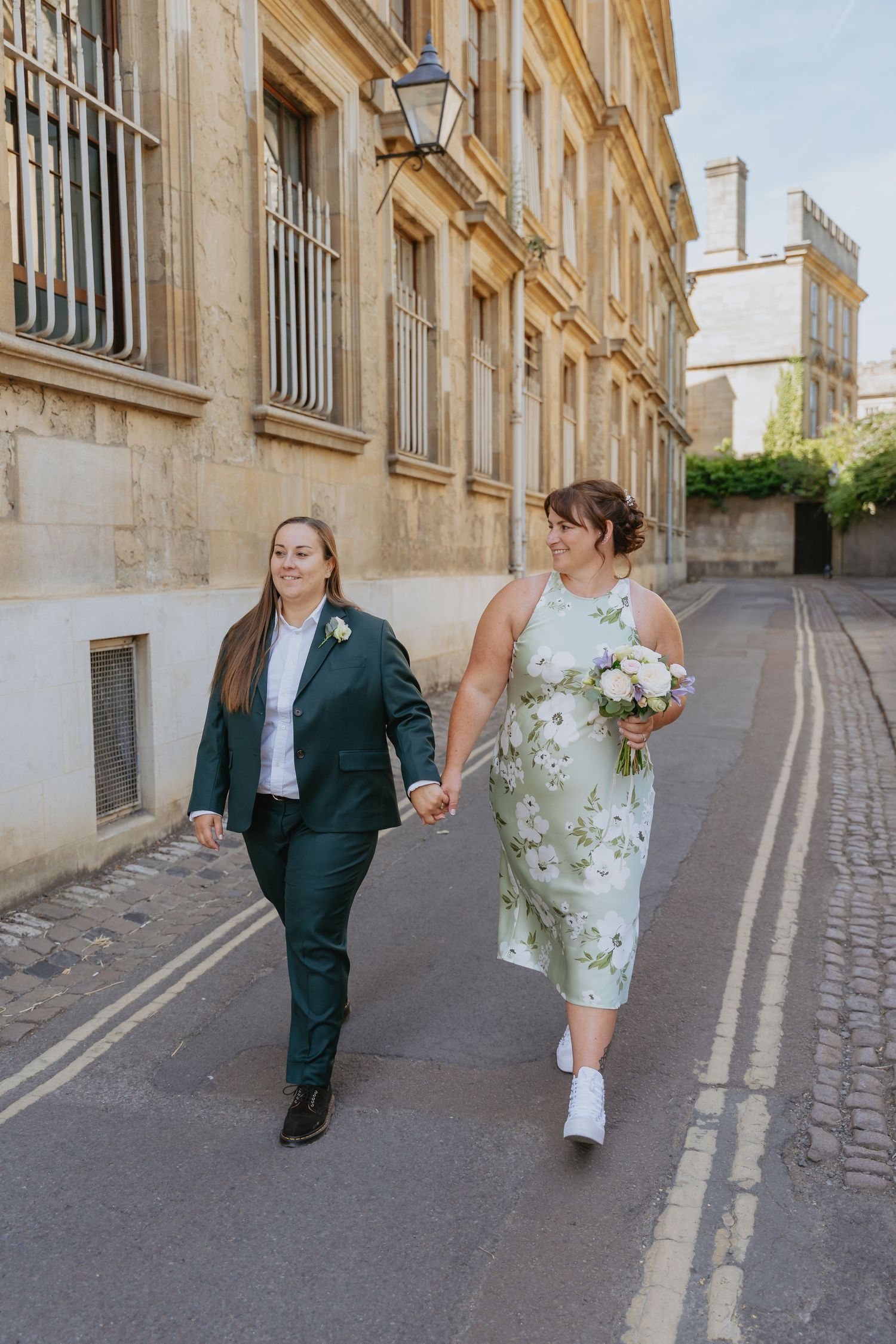 Two brides walk hand-in-hand down a sunny street, one in a green suit, the other in a floral dress holding a bouquet.