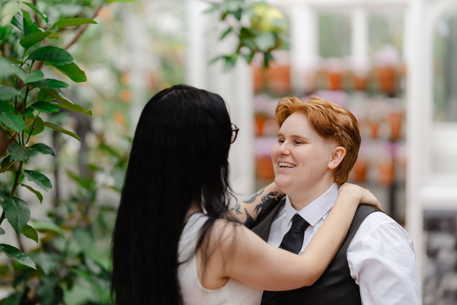 Two women sharing a joyful moment in a greenhouse, one in a vest and tie smiling warmly at the other.