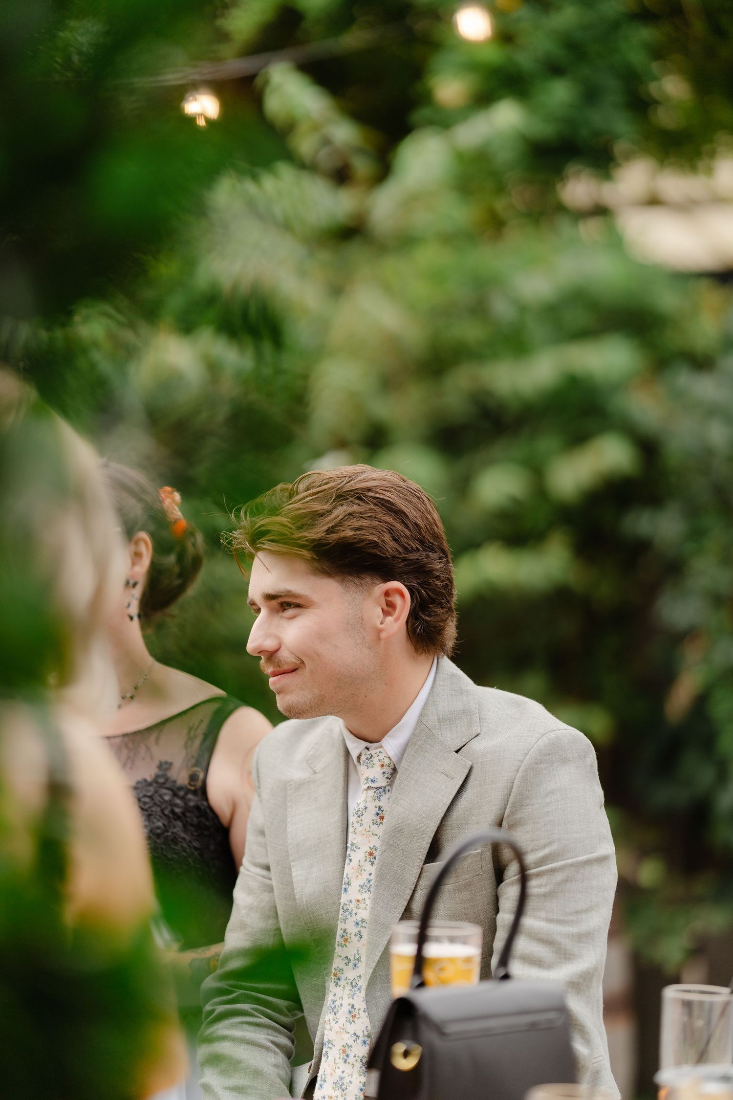 A smiling young man in a gray suit and floral tie sits at an outdoor garden dinner party surrounded by lush greenery.