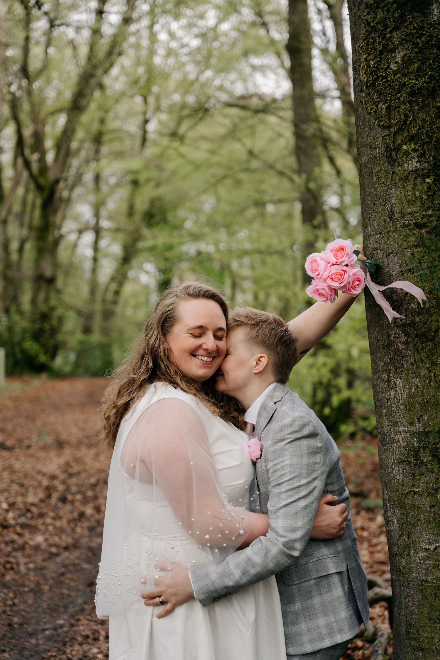 Couple in wedding attire sharing a tender kiss in a woodland setting, bride holding pink roses bouquet.
