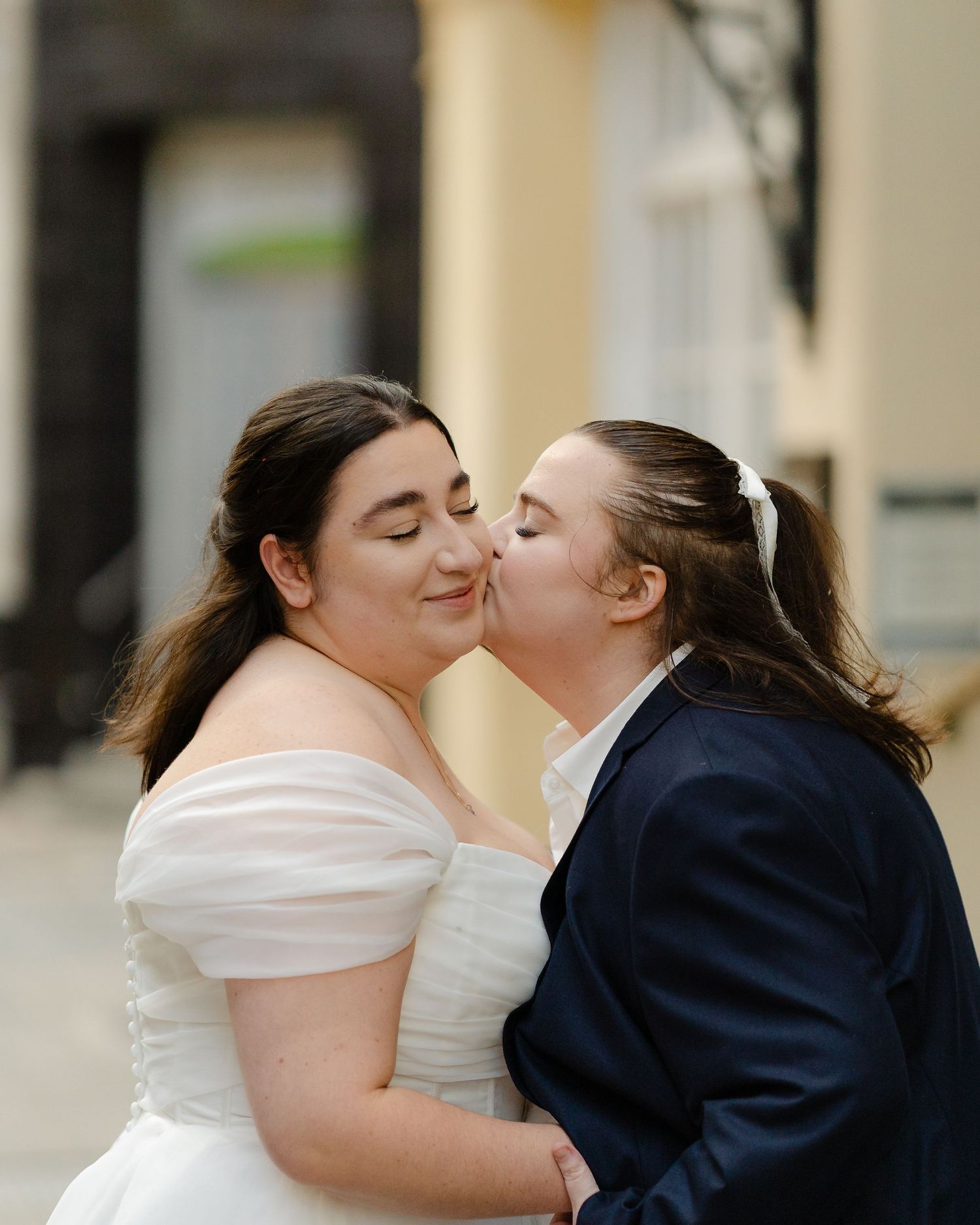 Two brides sharing a kiss, one in a white off-shoulder gown and one in a navy suit, at an outdoor wedding.