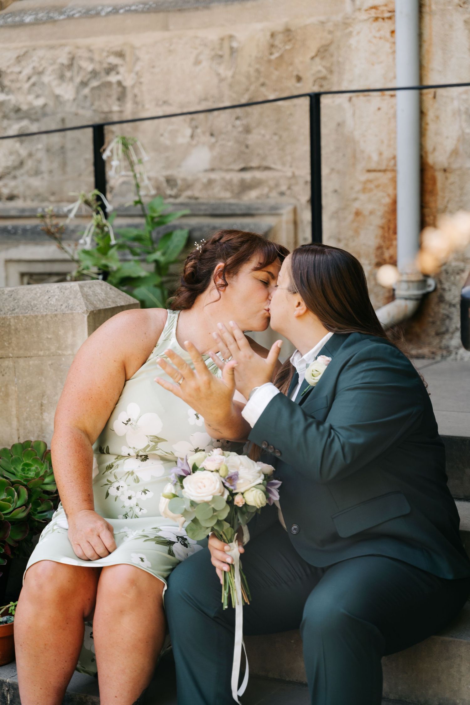 Two brides share a kiss on stone steps, one in a floral dress holding a bouquet, the other in a dark suit.