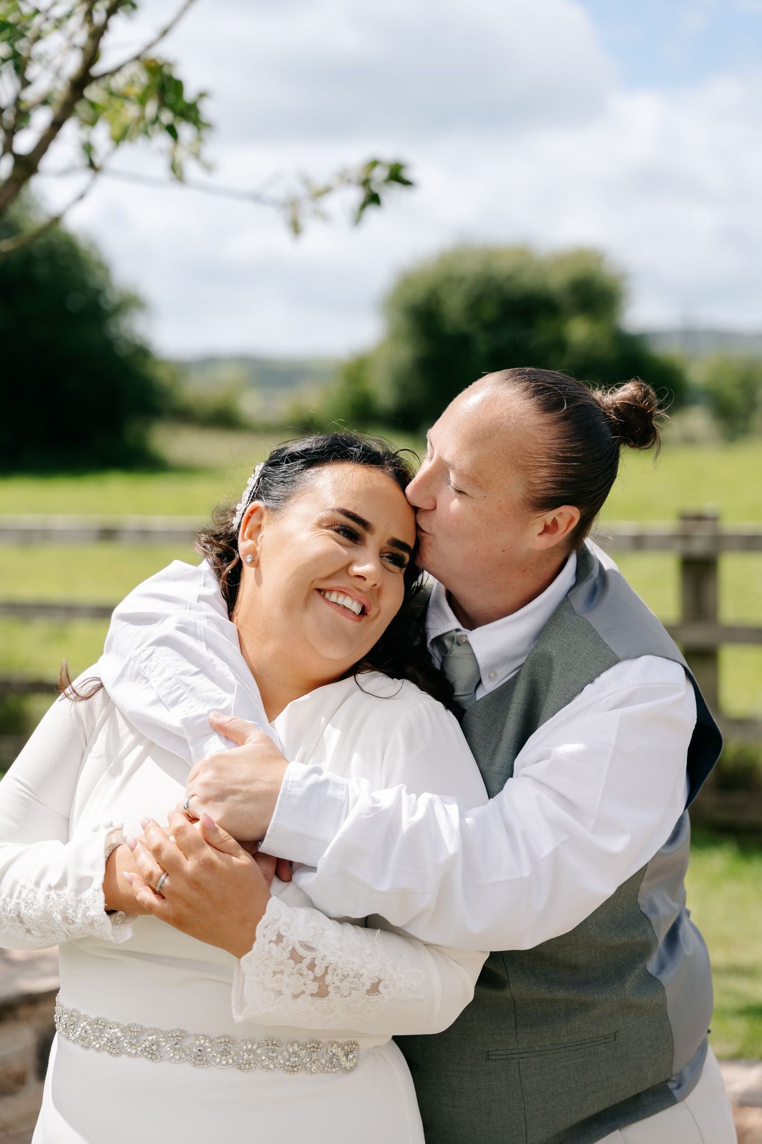A groom kisses his smiling bride on the cheek while embracing her outdoors at a sunny countryside wedding venue.