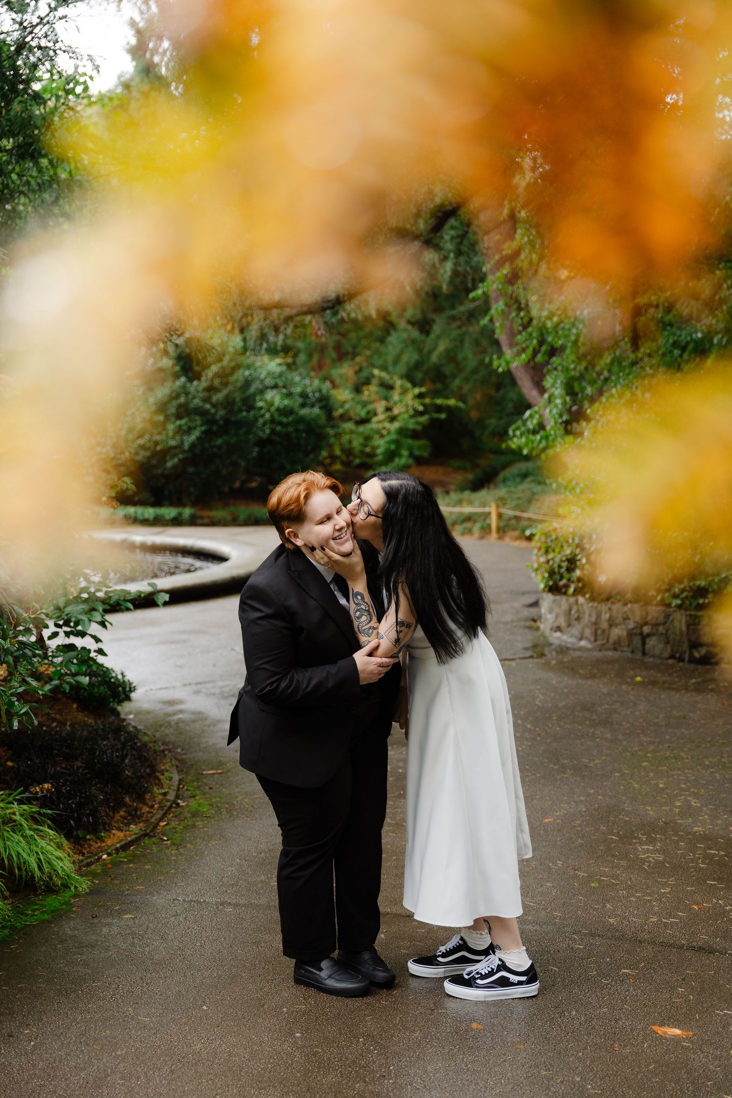 Two brides share a kiss in an autumn garden, one in a black suit and one in a white dress with Vans sneakers.