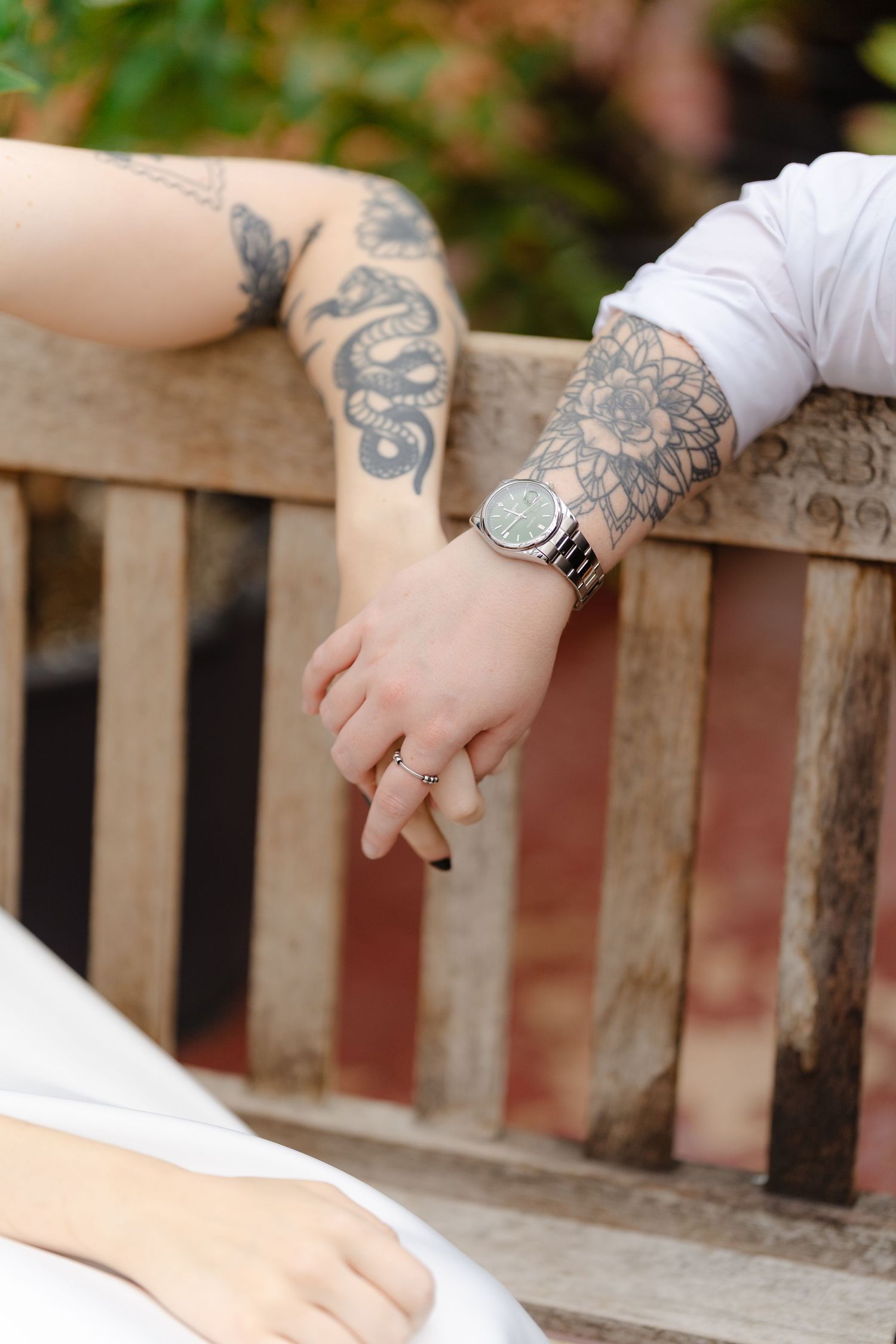 Tattooed couple holding hands on a wooden bench, man wearing a silver watch and woman with a ring.