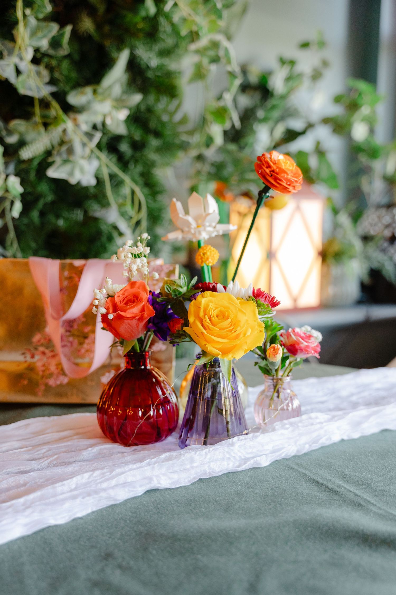 Colorful floral arrangements in red, purple, and clear glass vases on a white linen table runner with green foliage backdrop.