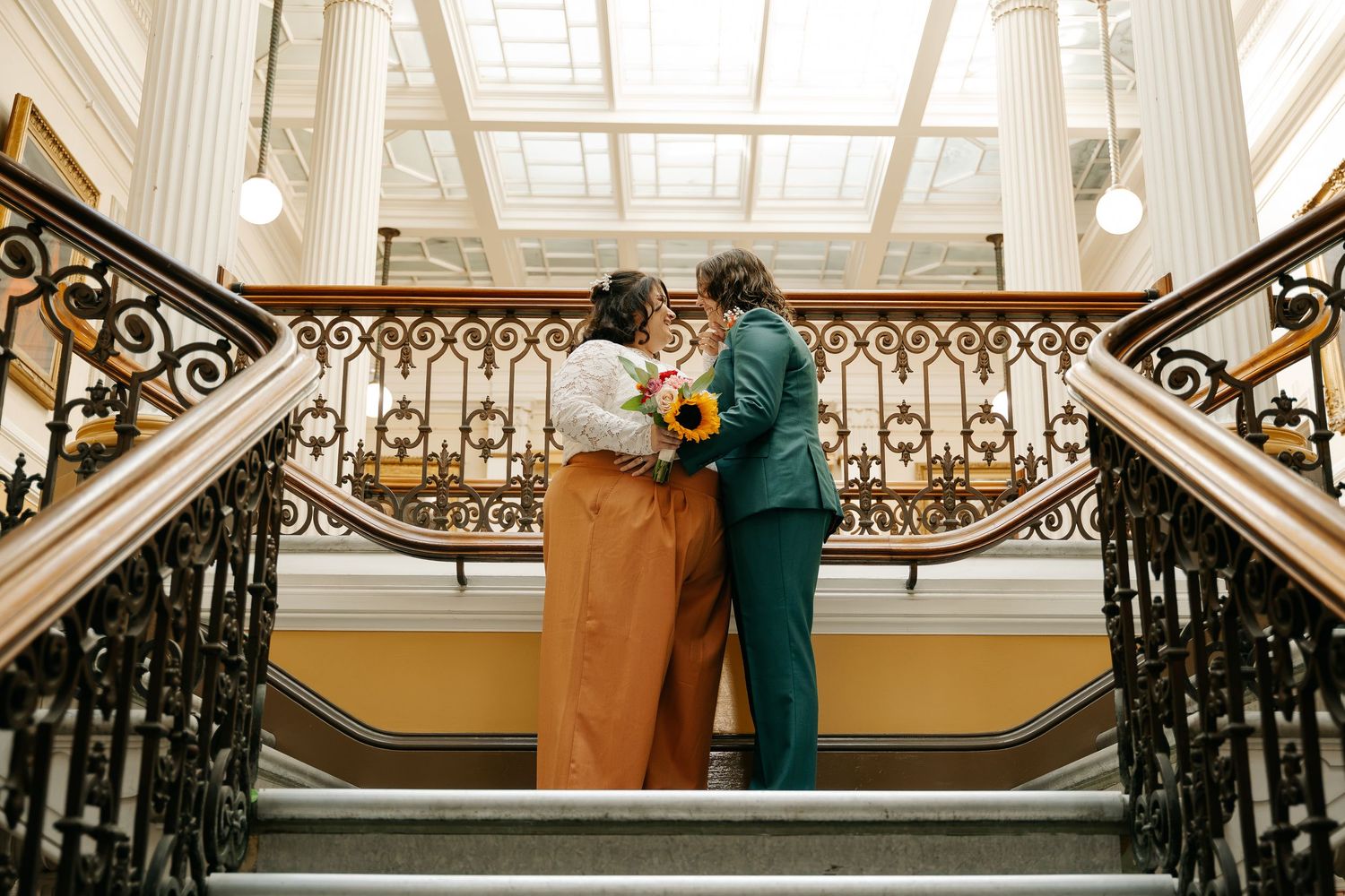 Couple embracing on grand staircase holding sunflowers, wearing teal suit and orange skirt at elegant wedding venue.