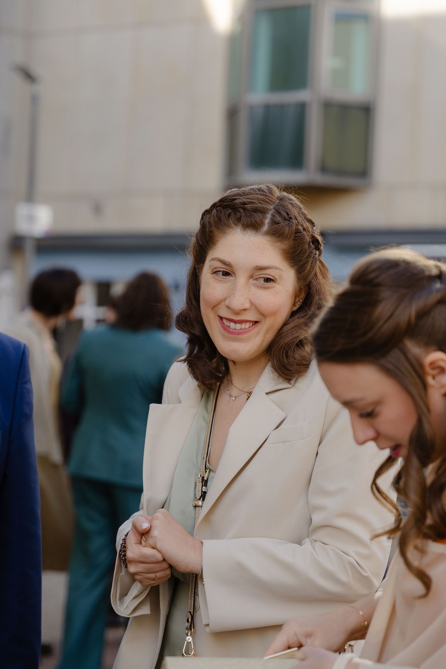 Smiling woman in beige blazer holding a purse at an outdoor social gathering with people in background.