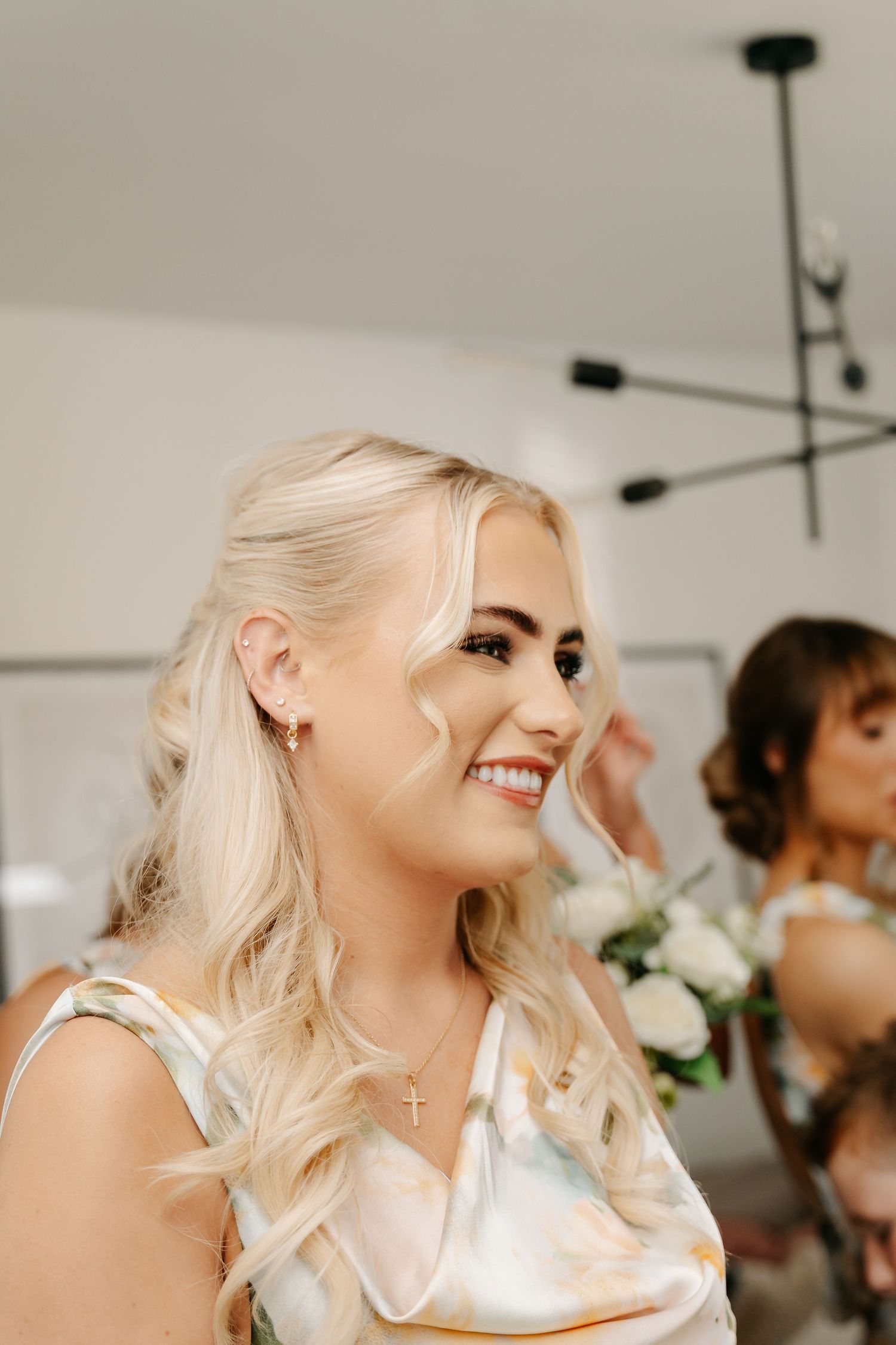 Smiling blonde bridesmaid wearing a cross necklace and satin dress, with white roses visible in the background.