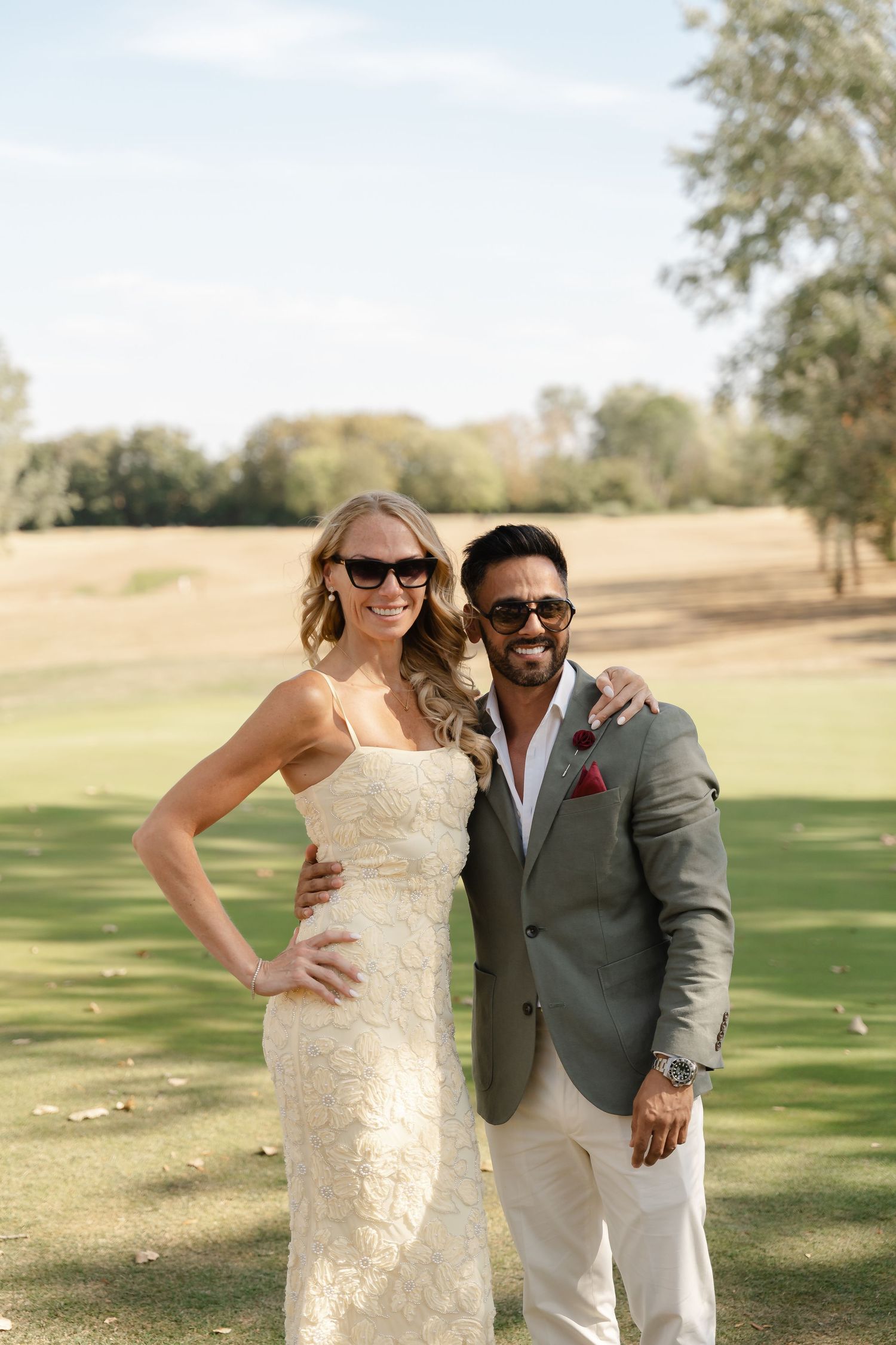 Smiling bride in lace gown and groom in gray suit with sunglasses pose together on a sunny golf course.