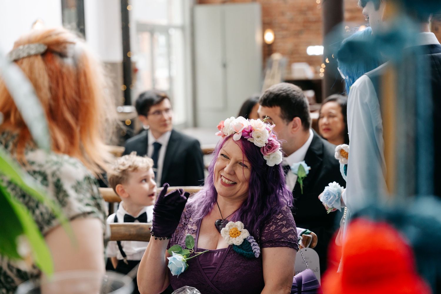Woman with purple hair and floral crown laughing joyfully at a wedding ceremony surrounded by guests.