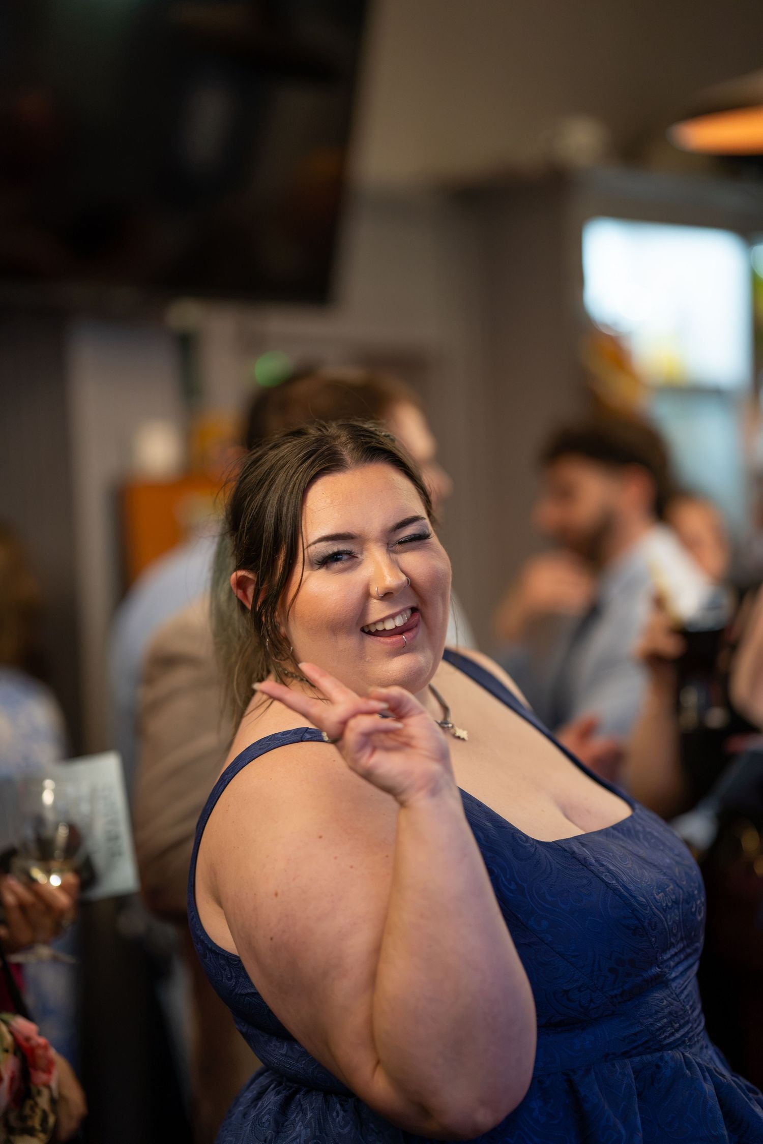 A smiling woman in a navy blue dress poses playfully at an indoor social gathering or wedding reception.