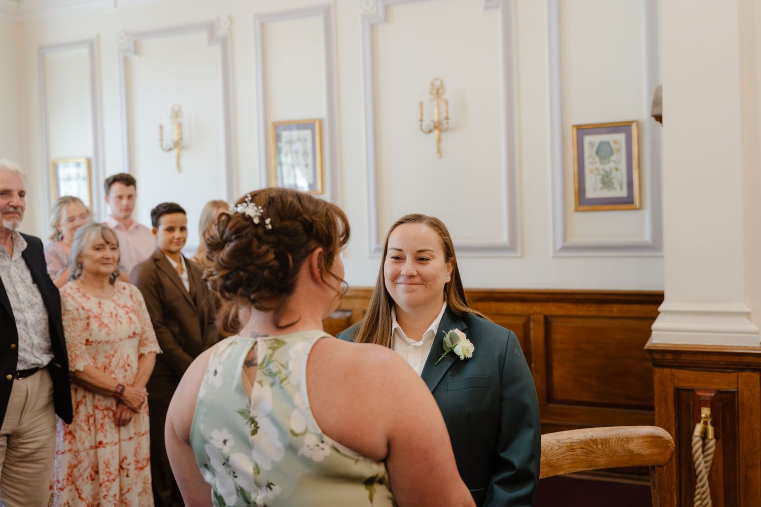 Two brides share a joyful moment at their wedding ceremony, surrounded by smiling guests in an elegant venue.