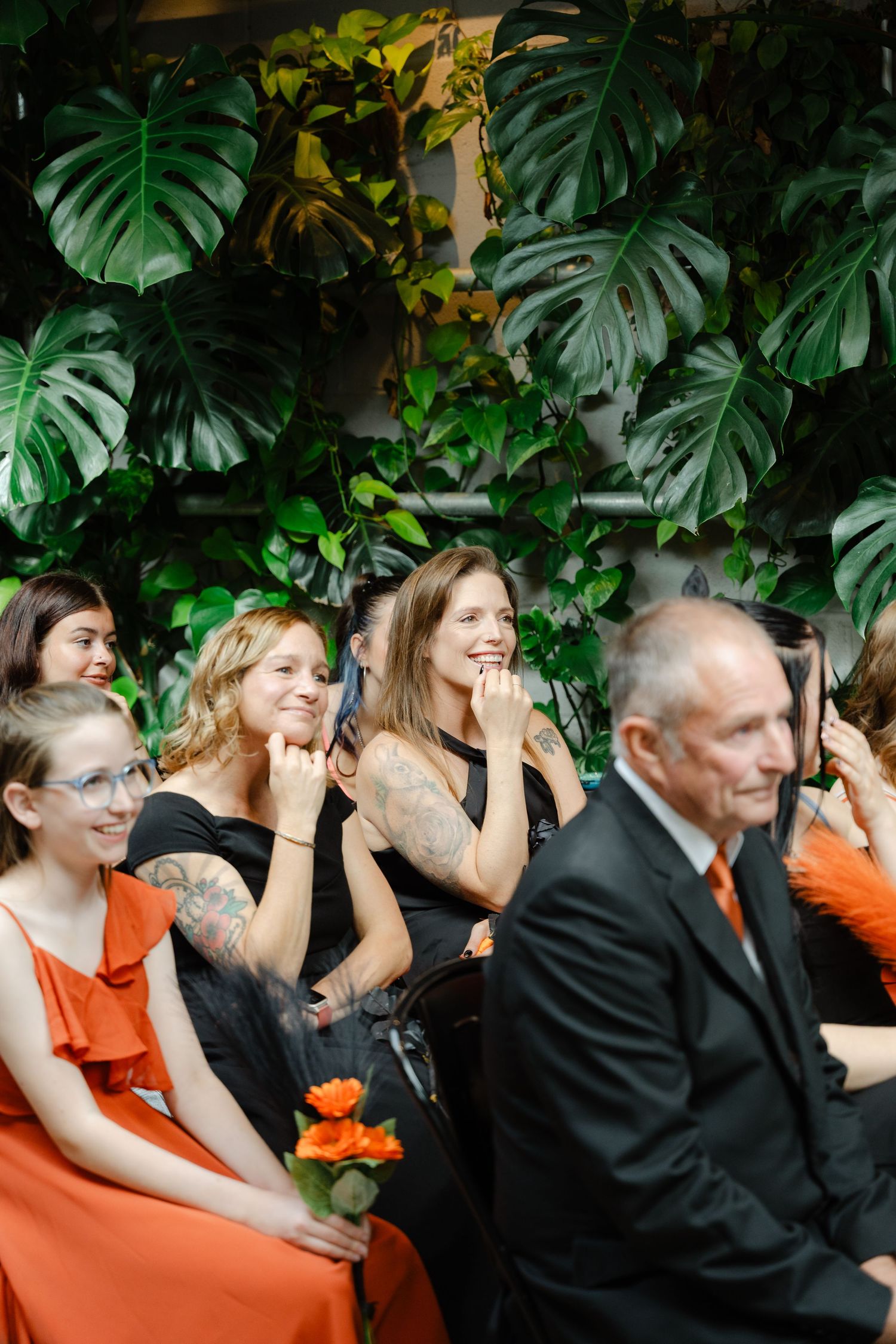 Smiling wedding guests in black and orange attire seated before lush tropical monstera leaf backdrop.