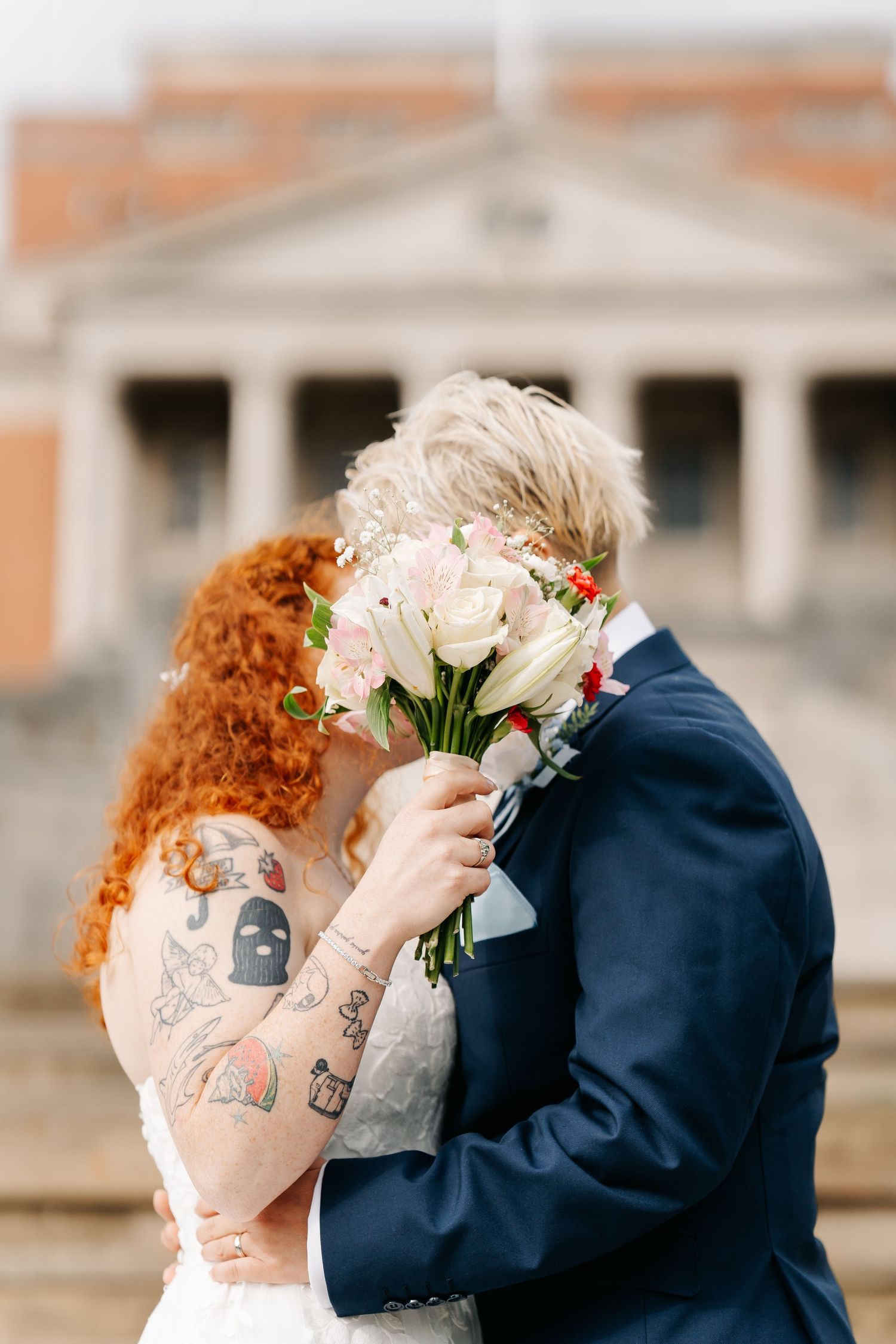 Bride with red curly hair and tattoos kisses groom in navy suit, hiding faces behind wedding bouquet outdoors.
