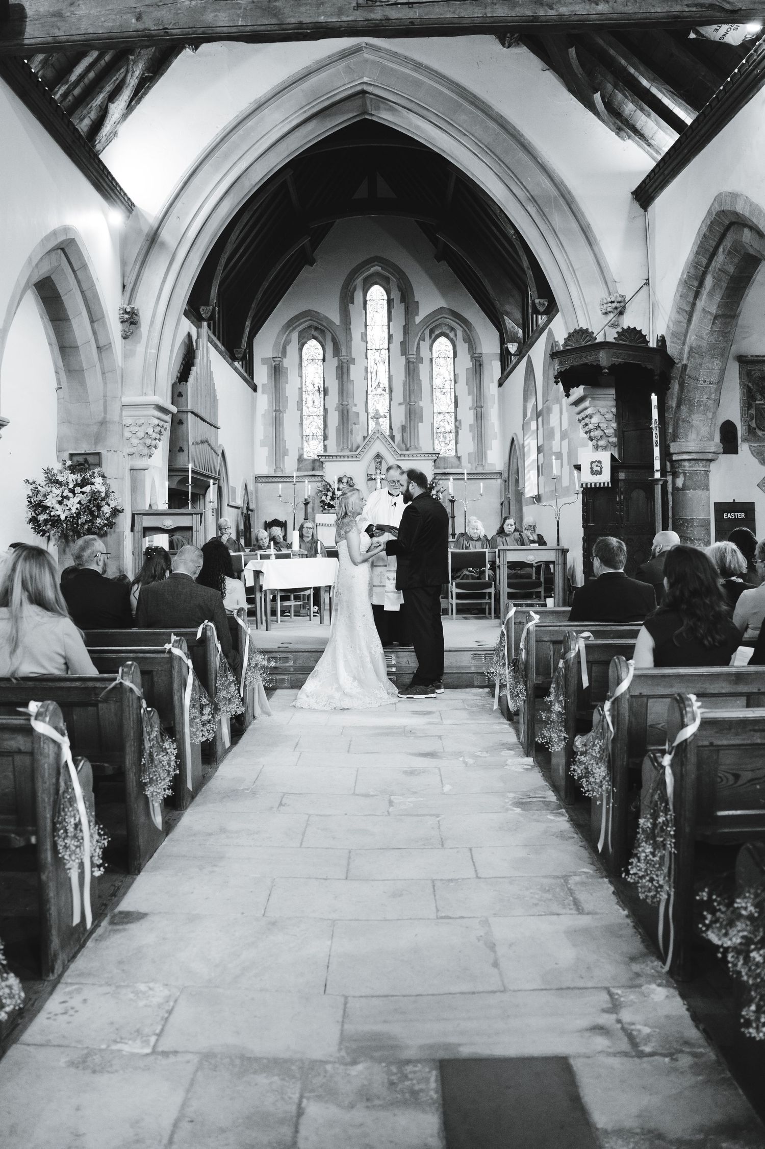 Black and white photo of a wedding ceremony inside a traditional stone church with guests seated in wooden pews.