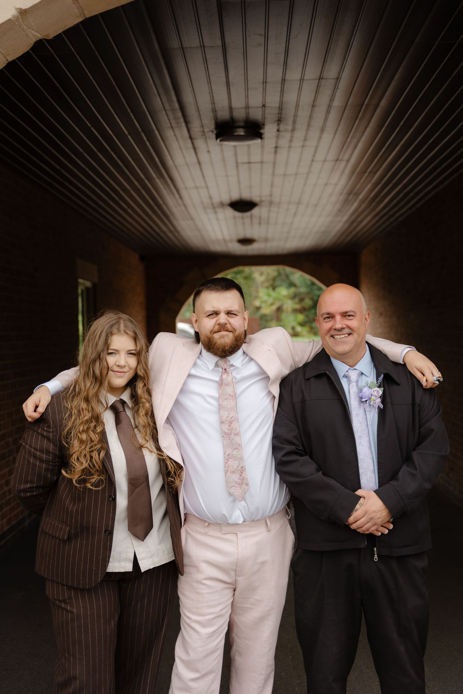 Three formally dressed people pose together in a brick archway, with the center man in a light pink suit embracing the others.