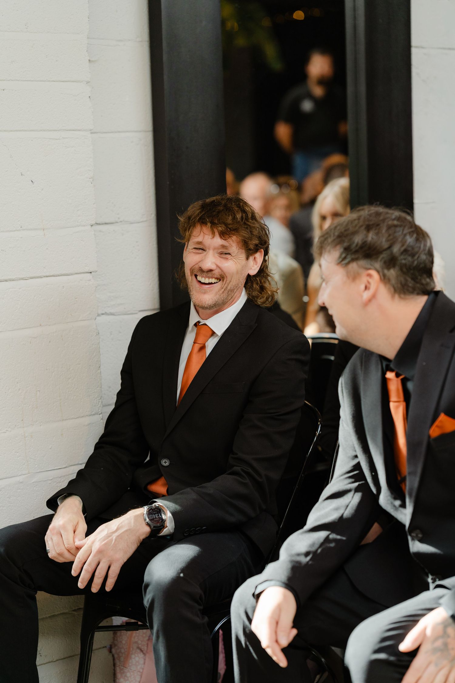 Two men in black suits with orange ties laughing together at a formal event or wedding ceremony.