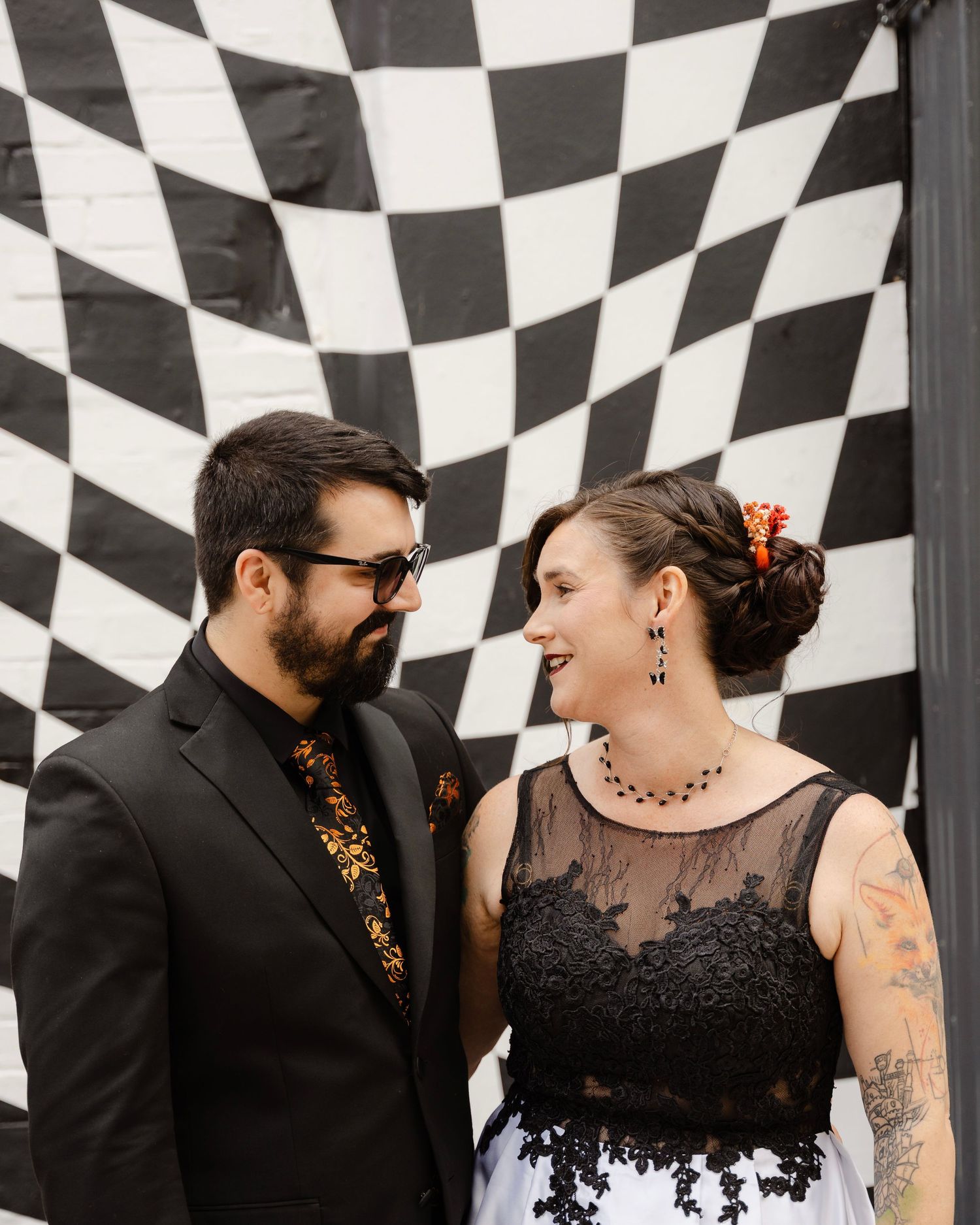 Couple in black wedding attire smiling at each other in front of a bold black and white checkered backdrop.