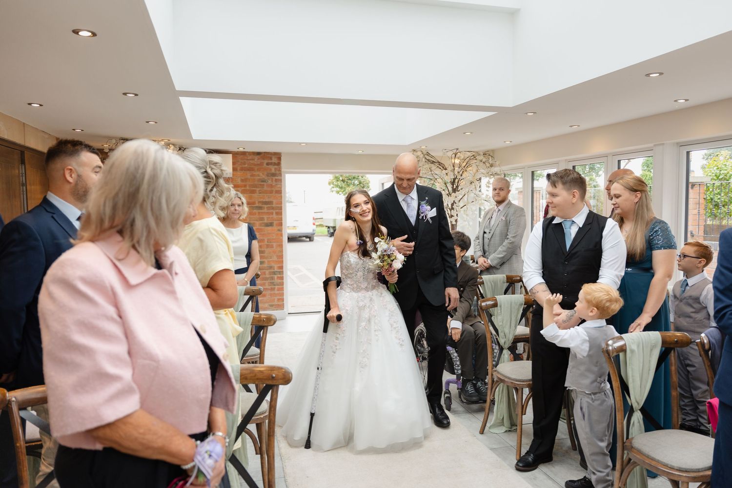 Bride walking down the aisle with escort at elegant indoor wedding ceremony, guests smiling in bright venue.