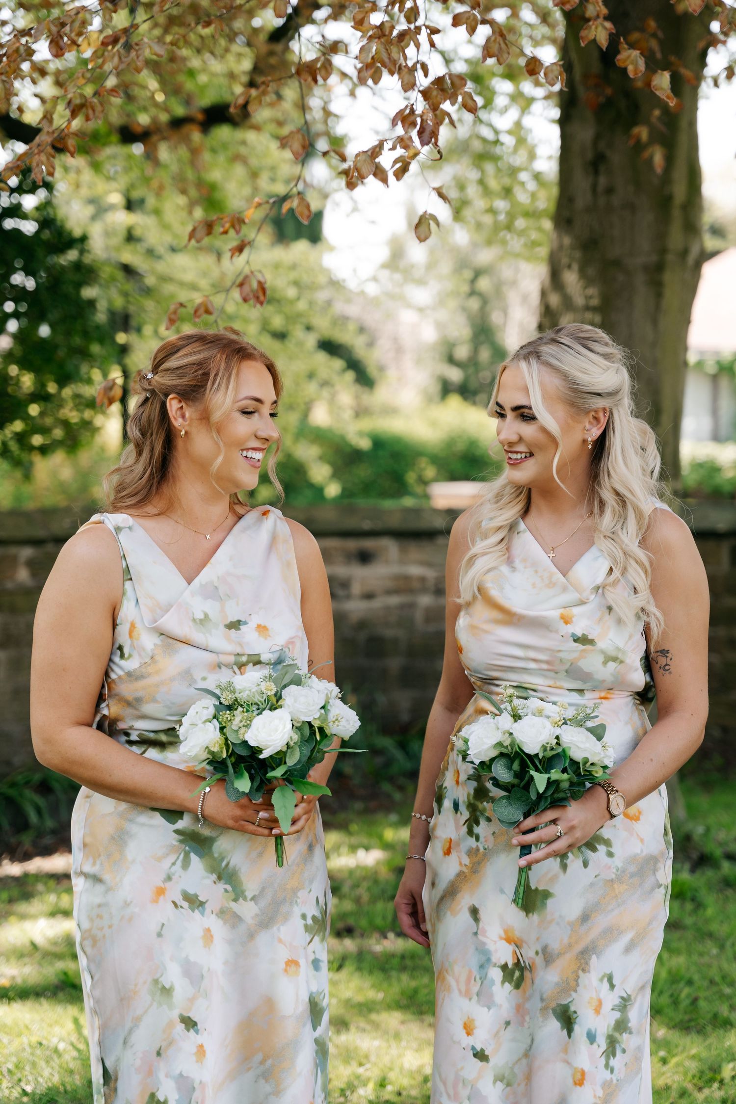 Two bridesmaids in matching floral dresses holding white bouquets, smiling outdoors under a tree.