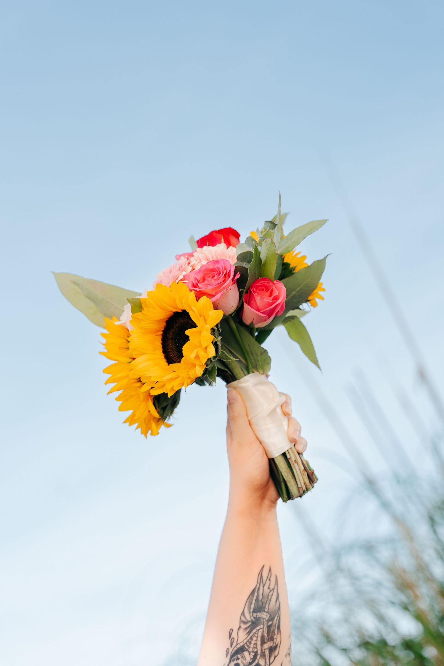 Tattooed hand holding a vibrant bouquet of sunflowers and pink roses against a clear blue sky.