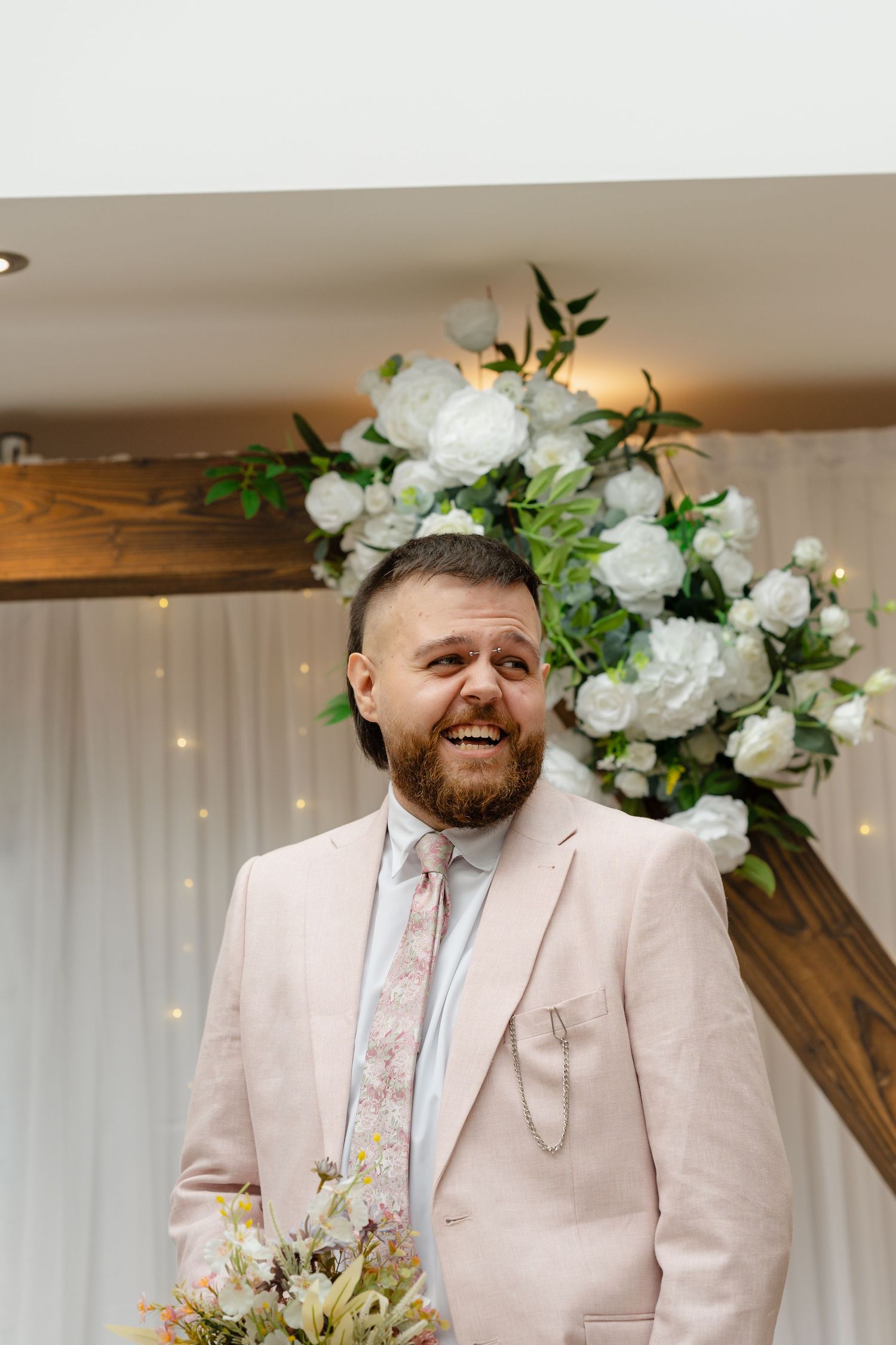 Smiling bearded groom in pink suit stands before white floral arch at wedding ceremony.