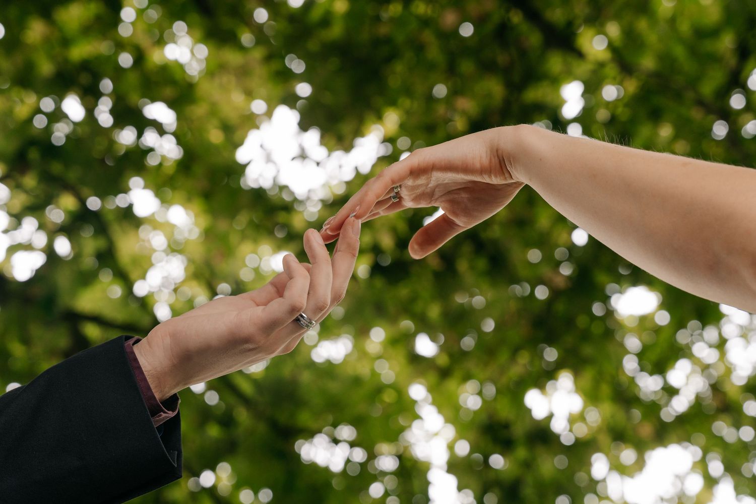 Two hands gently touching fingertips against a lush green bokeh background, symbolizing connection and romance.