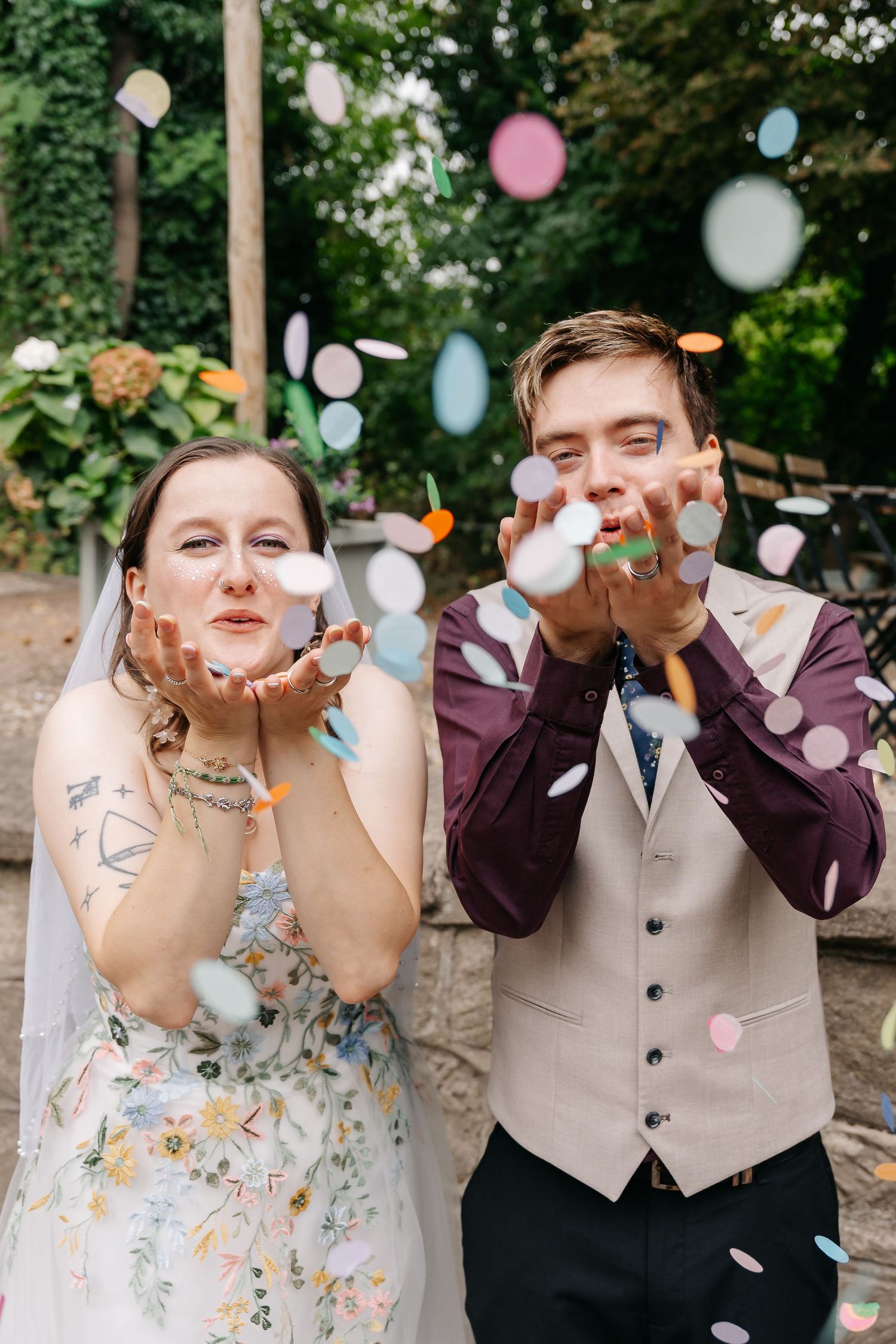 A bride and groom blow colorful confetti from their hands outdoors on their wedding day.