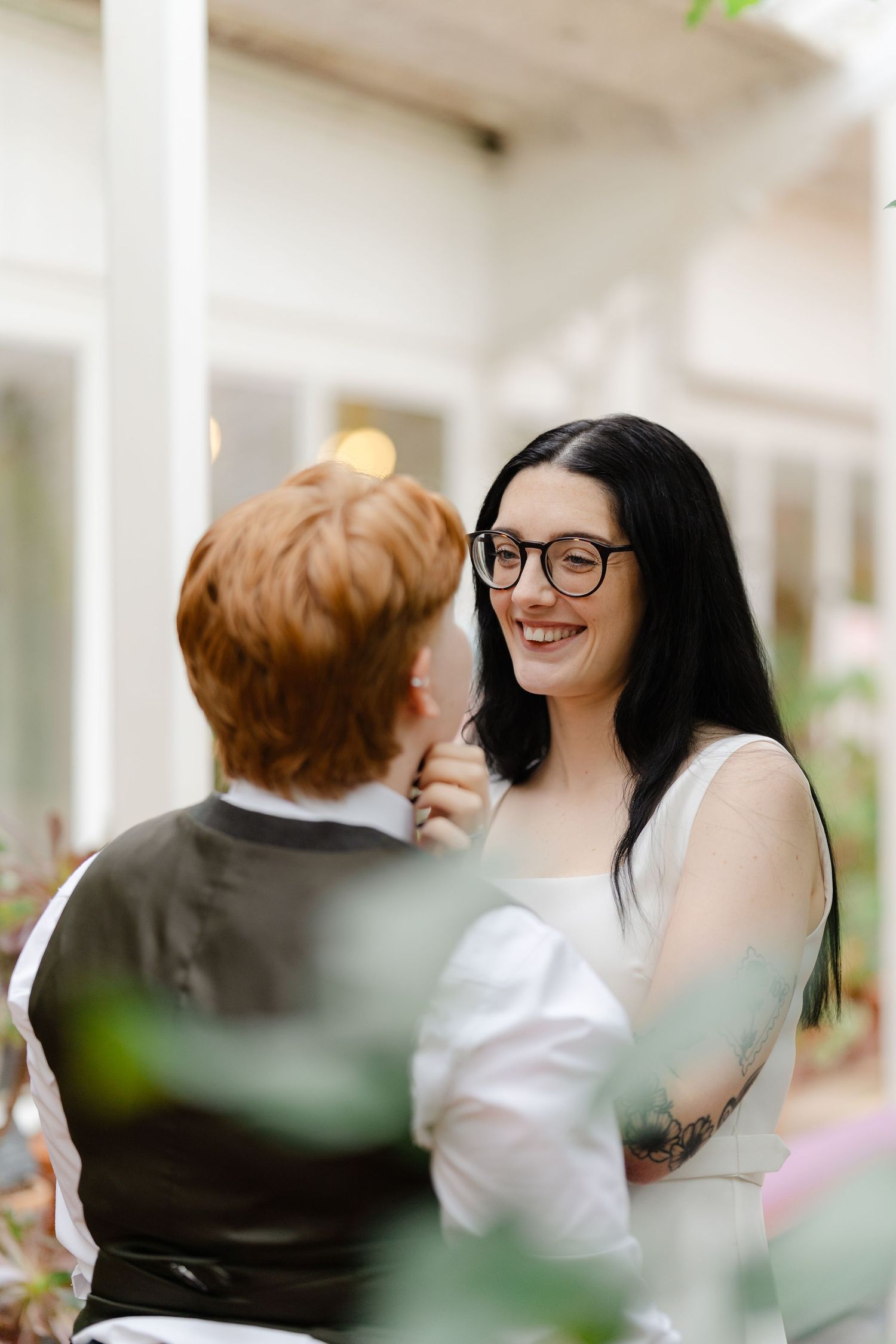 A smiling dark-haired bride in glasses shares a tender moment with a red-haired partner in a vest at an outdoor wedding.