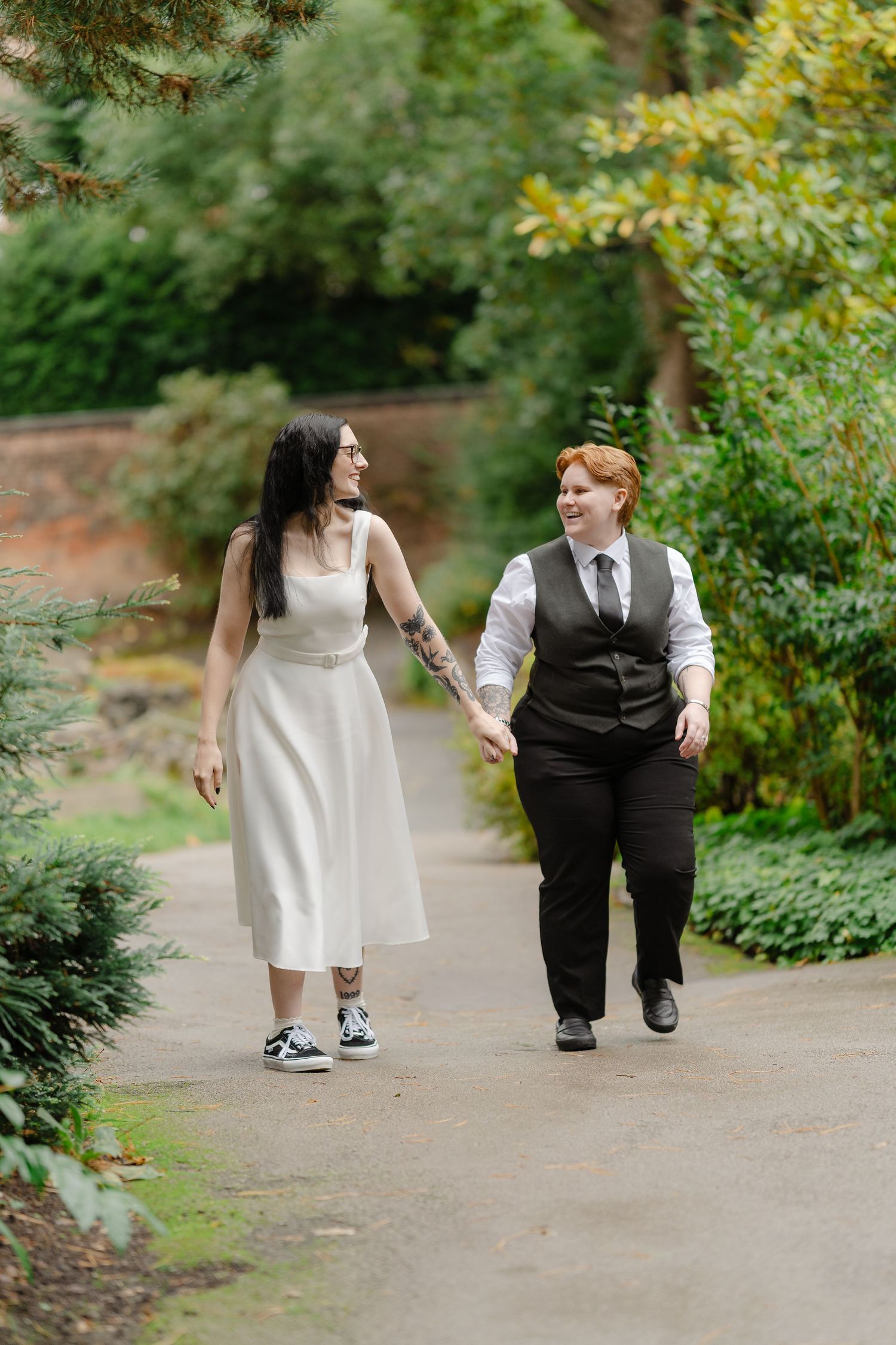 Two brides walk hand in hand through a lush garden, one in a white dress and one in a black vest and tie.