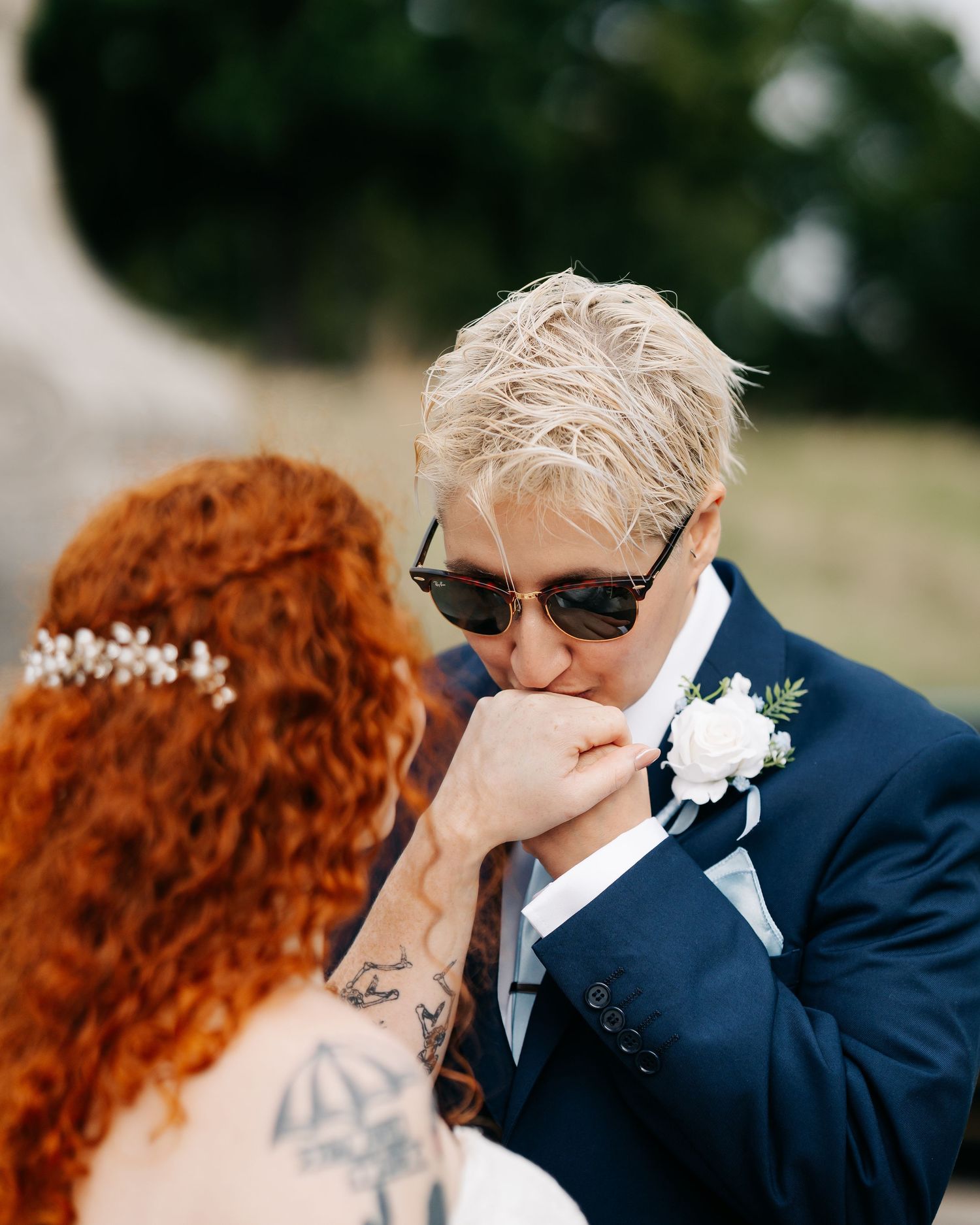 A person in a navy suit kisses the hand of a redhead bride wearing a floral hair piece at a wedding.